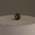 The Thick Pave Onyx Barrel Charm, featuring gold and black beads, rests on a textured light stone surface against a plain background.