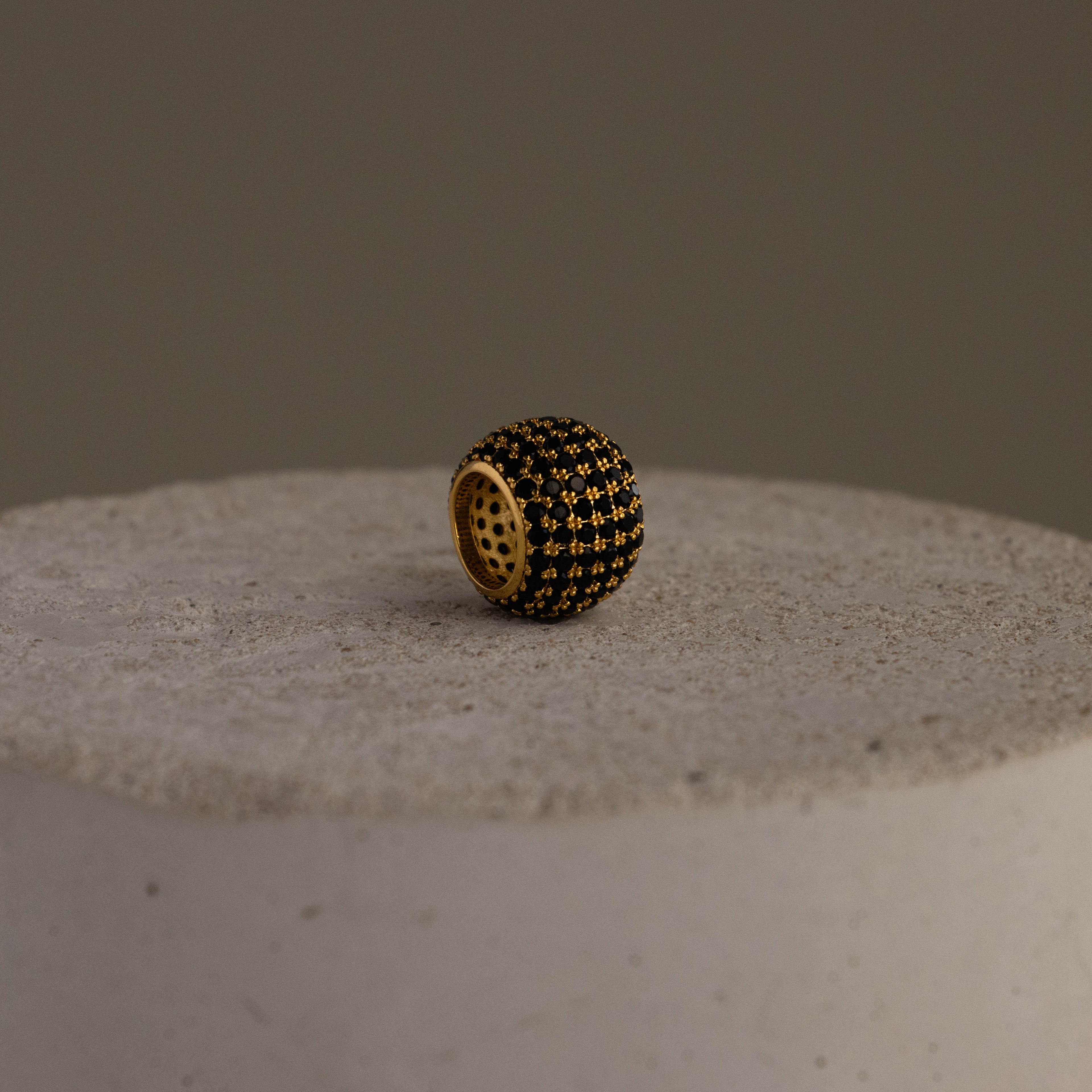 The Thick Pave Onyx Barrel Charm, featuring gold and black beads, rests on a textured light stone surface against a plain background.