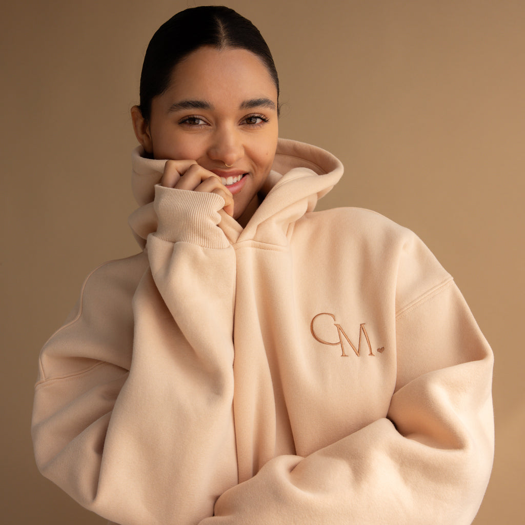 Smiling woman wears an Oversized CM Hoodie in beige, standing against a matching Caitlyn Minimalist background.