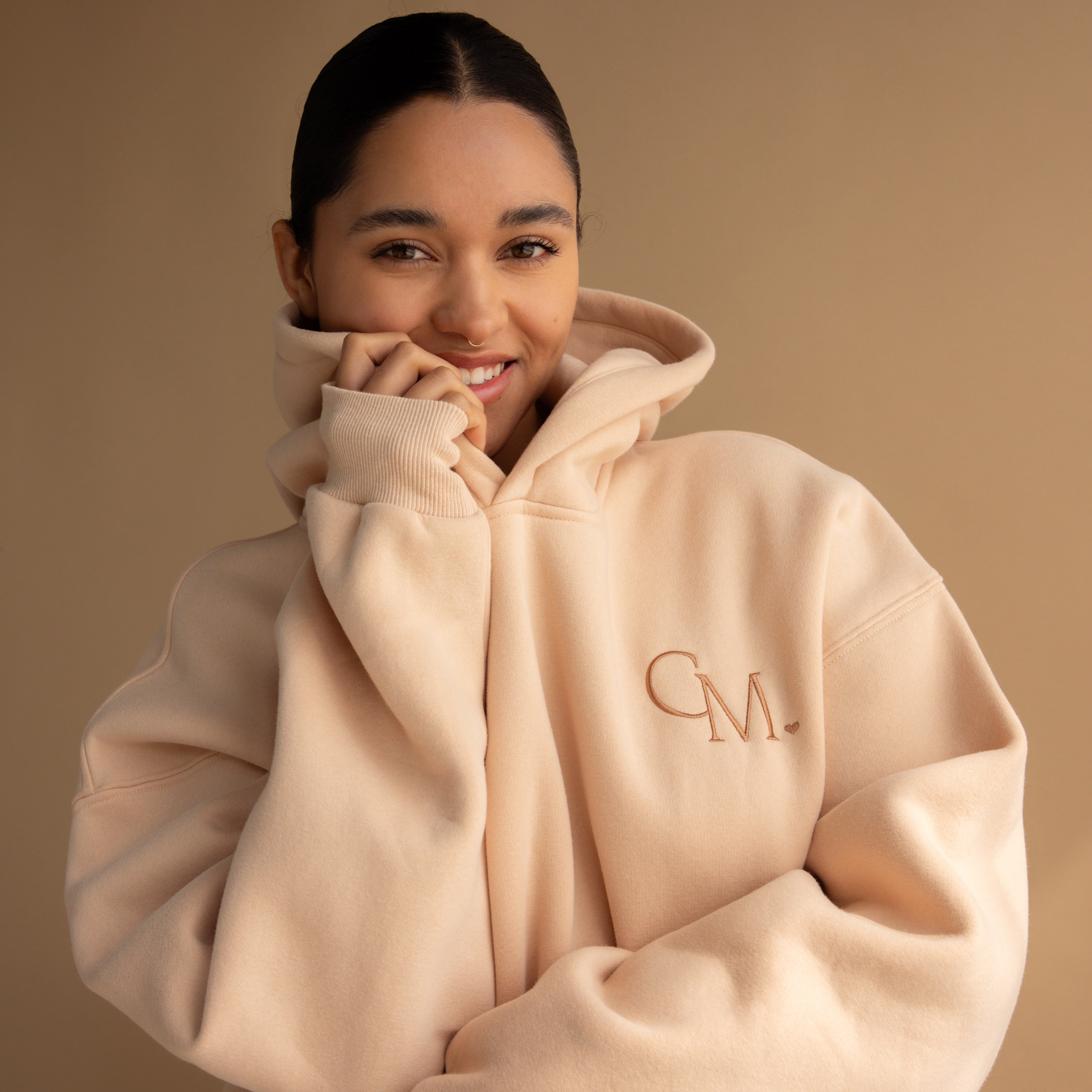 Smiling woman wears an Oversized CM Hoodie in beige, standing against a matching Caitlyn Minimalist background.