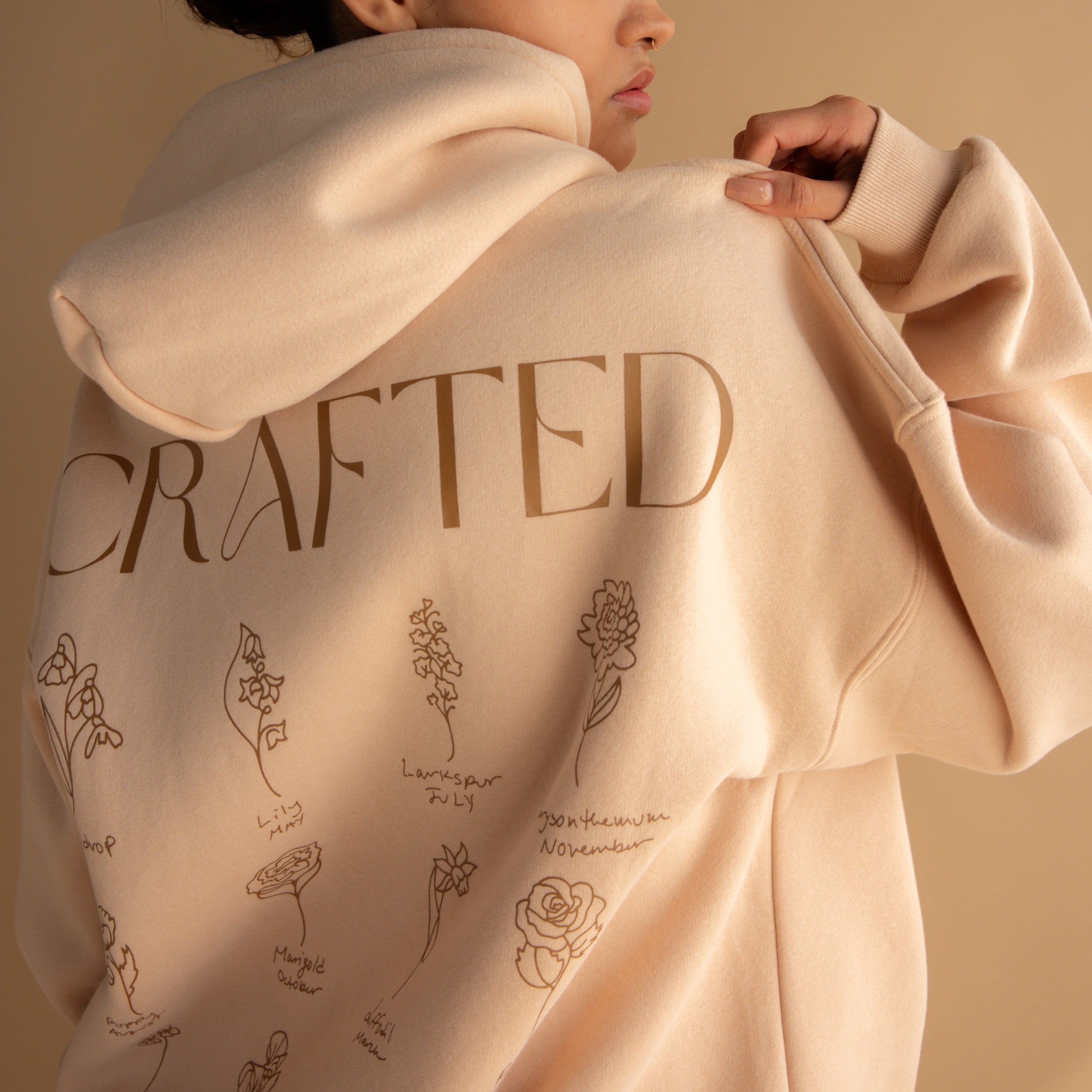 A person wearing an Oversized CM Hoodie in beige, featuring CRAFTED and flower prints on the back—ideal for fans of Caitlyn Minimalist style.