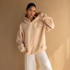 Woman wearing the Oversized CM Hoodie and white pants stands against a beige background, gazing at the camera—embodying the effortless Caitlyn Minimalist style.