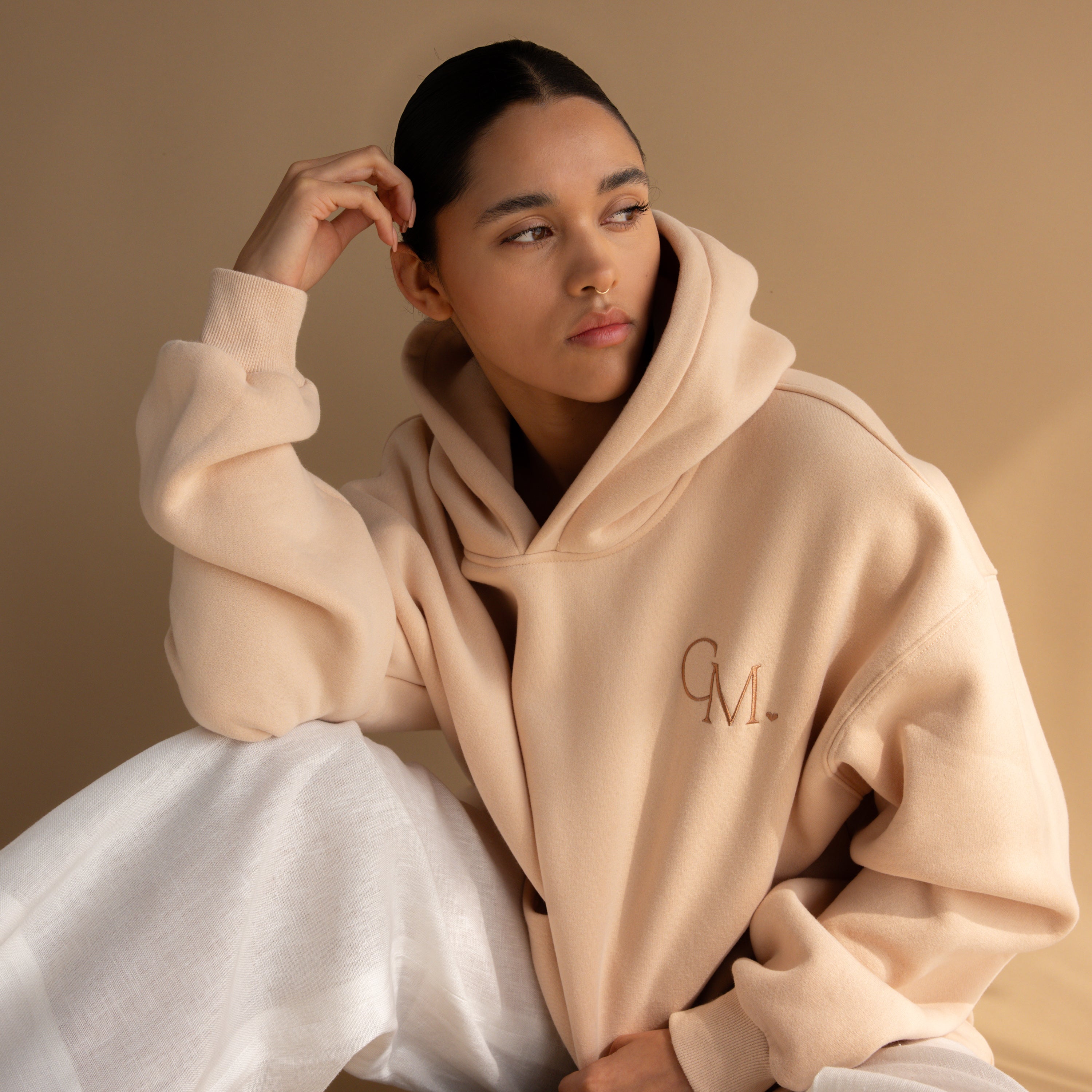 A woman wearing the Oversized CM Hoodie by Caitlyn Minimalist, featuring CM. embroidery, sits against a tan background and looks to the side.