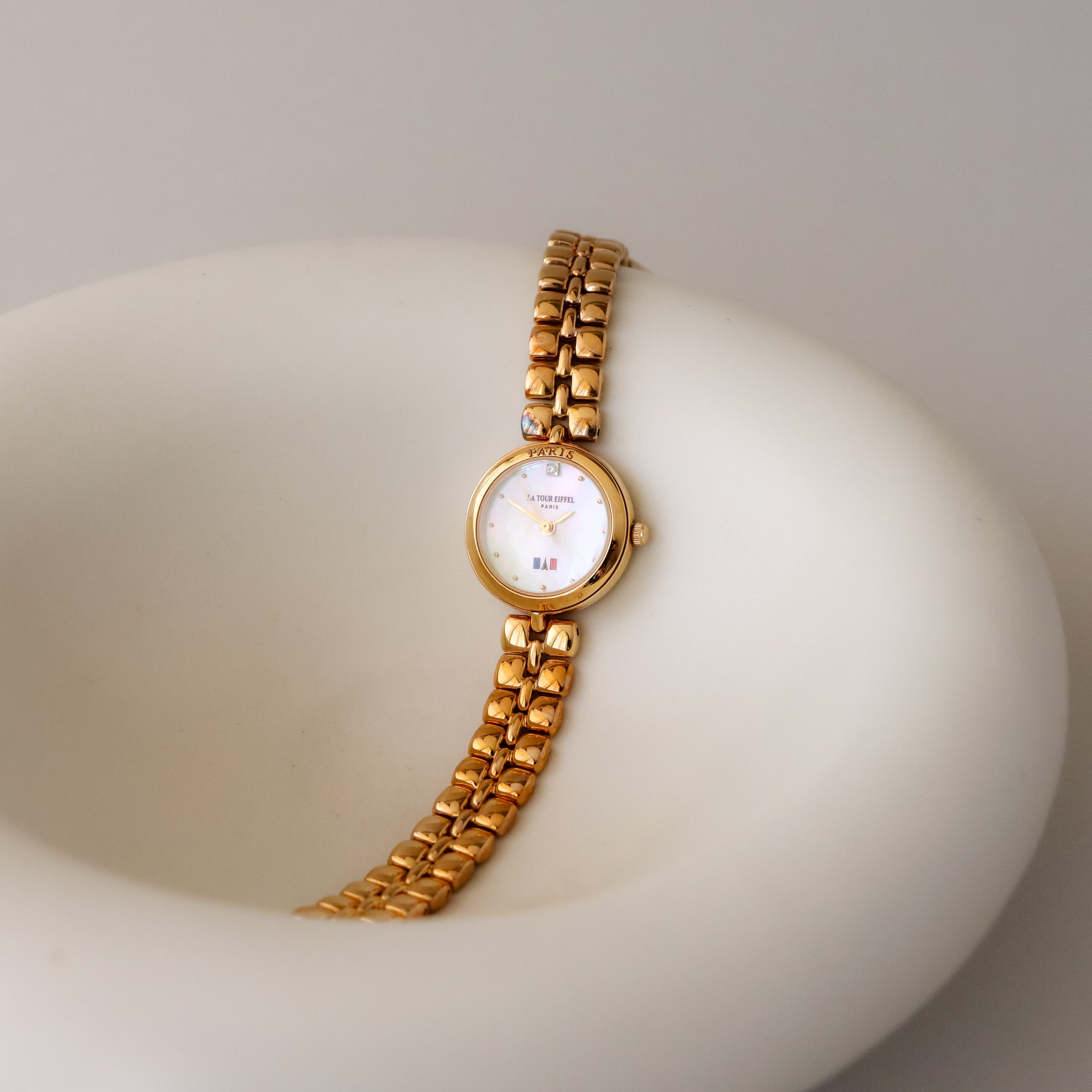 A Vintage La Tour Eiffel Round Gold Watch with a mother-of-pearl dial rests elegantly on a smooth white, bowl-shaped surface.