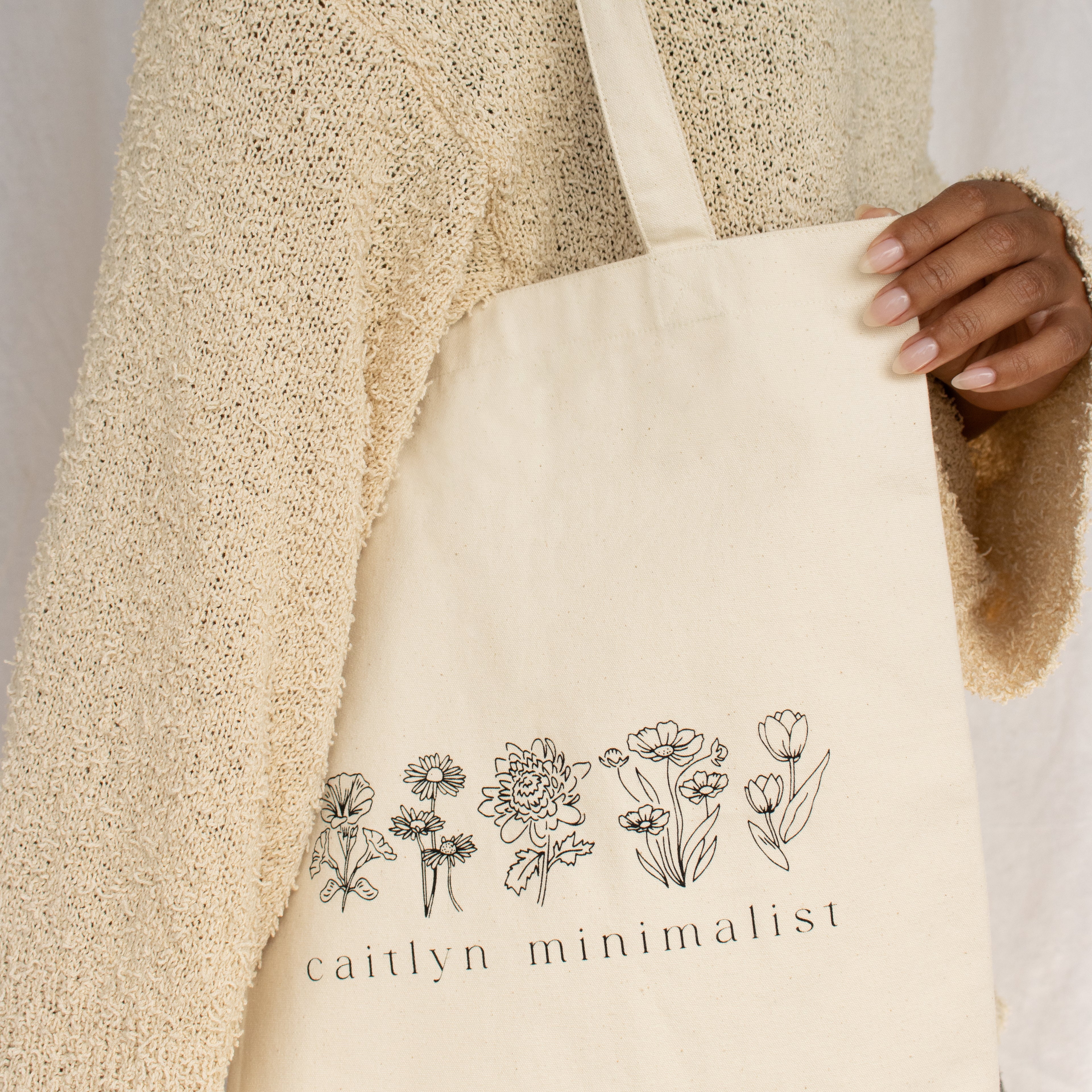A person carries the CM Tote Bag in cream, adorned with floral line art and the text "caitlyn minimalist.