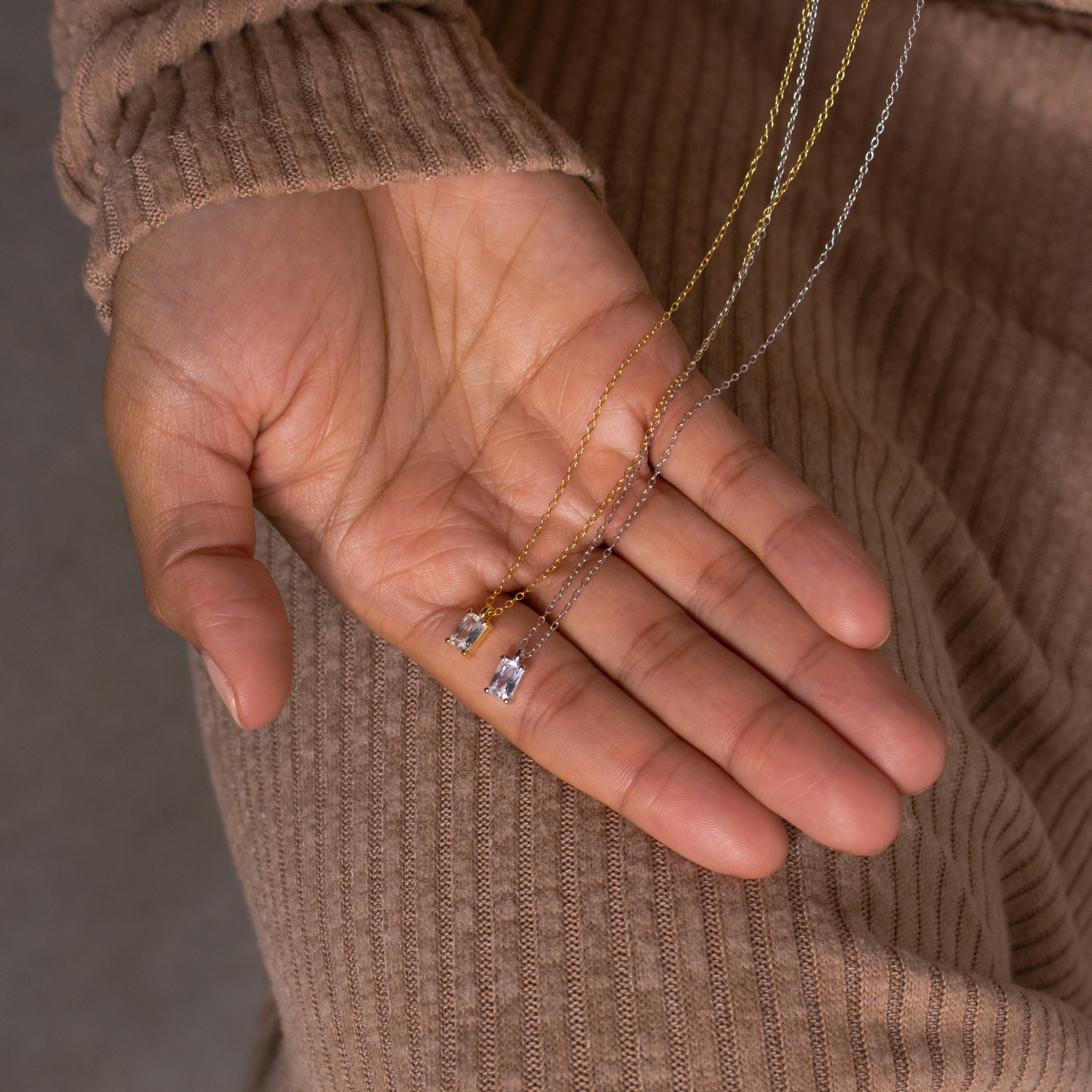 A hand holds two luxury necklaces with rectangular gemstone pendants, including the Sage Diamond Necklace, over a ribbed brown sleeve.
