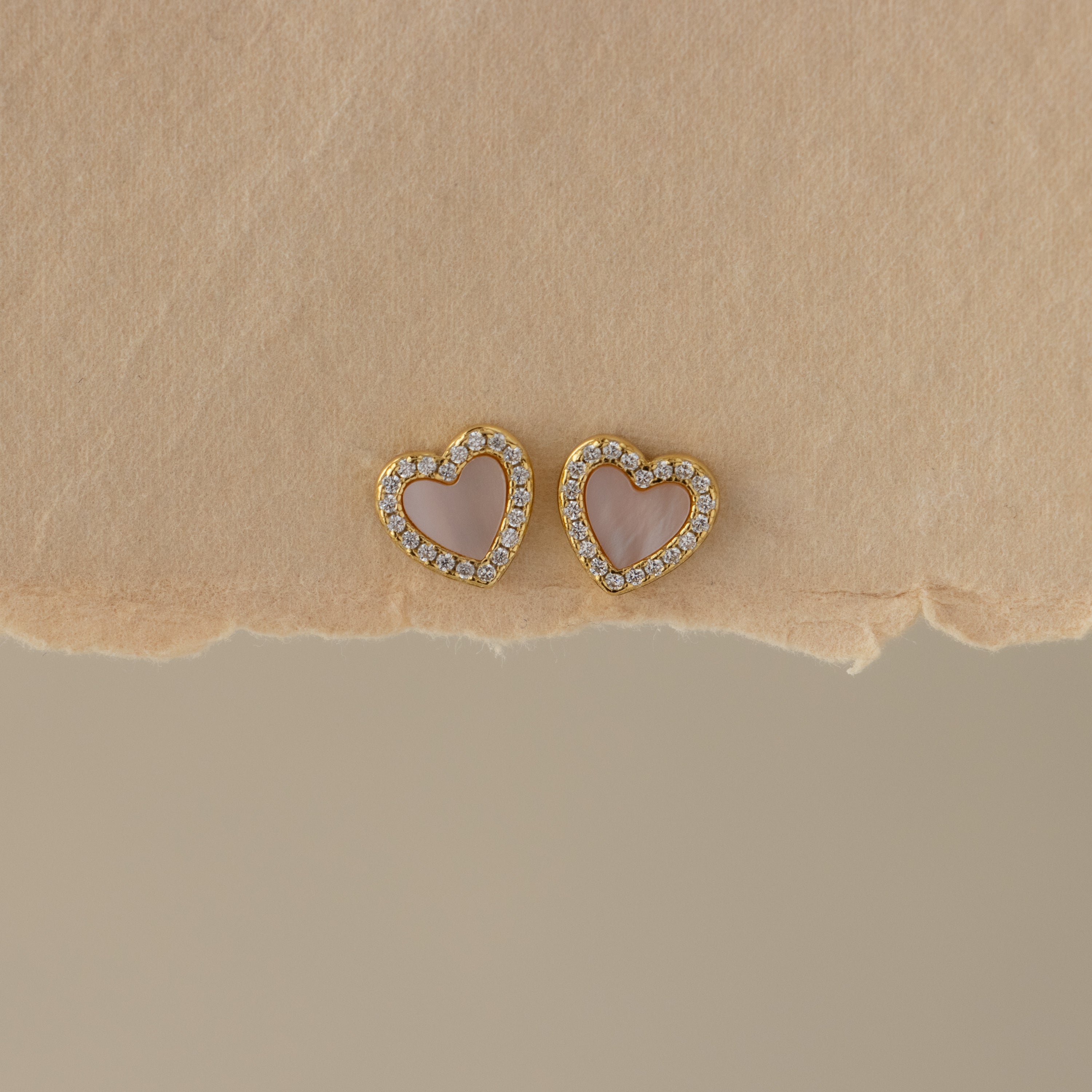 Pink Pave Heart Studs—gold heart-shaped earrings with pink centers and sparkling crystals—are displayed on textured beige paper.