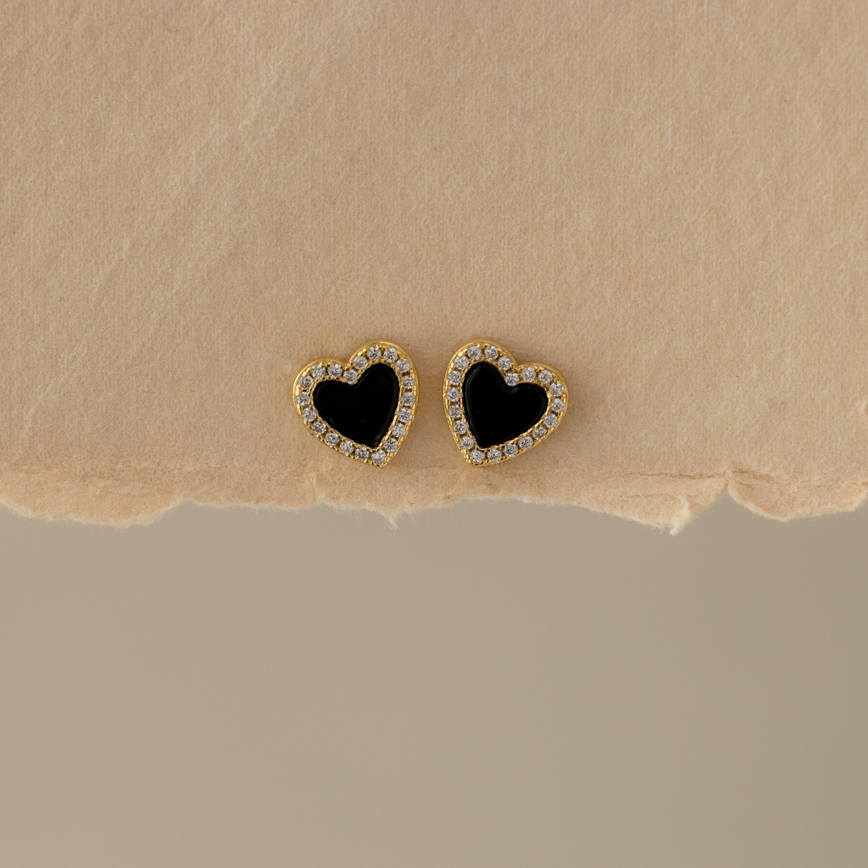 Black Pave Heart Studs: Gold heart-shaped earrings with black centers and clear stones, shown on a beige textured surface.
