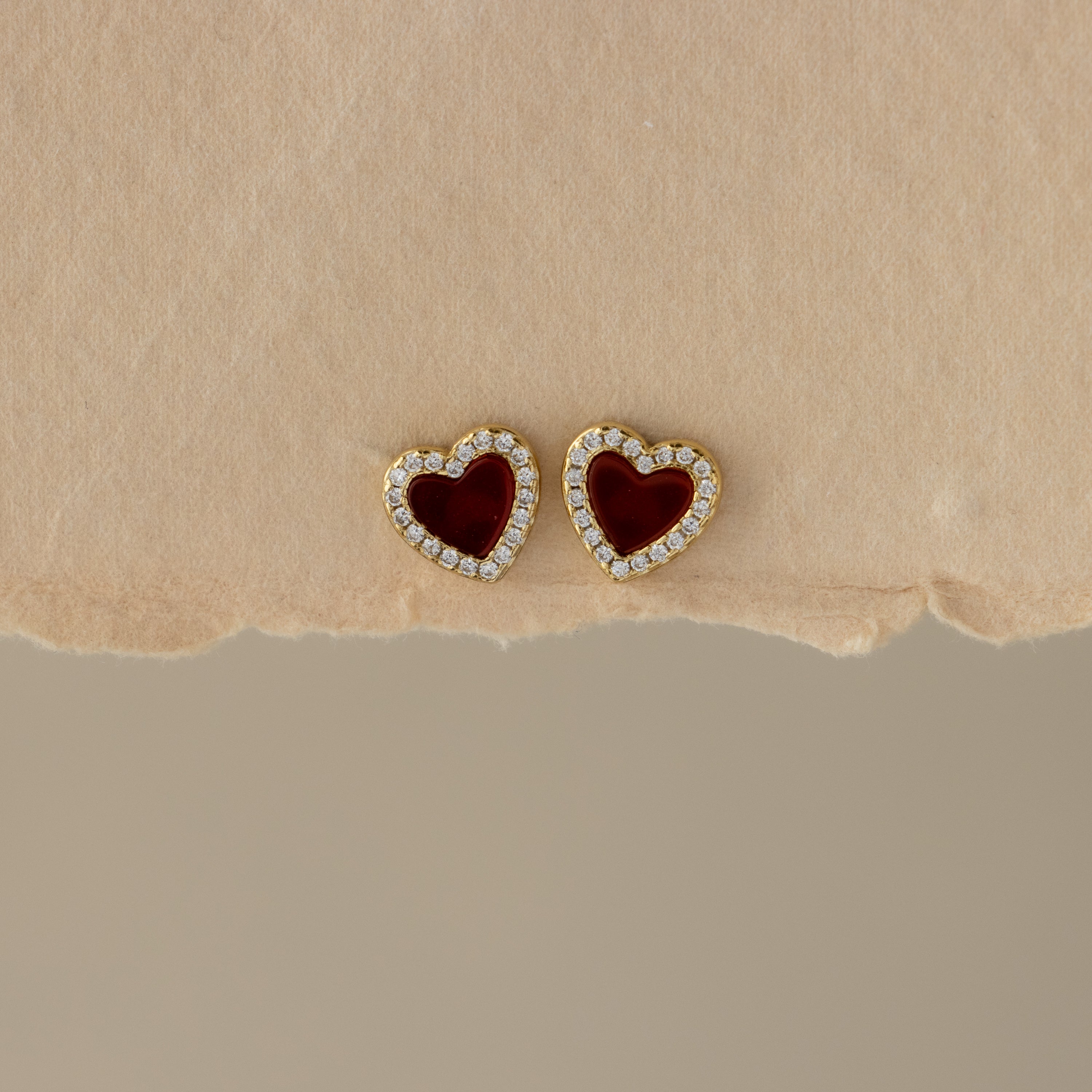 Red Agate Pave Heart Studs feature gold heart-shaped earrings with red centers and small clear stones outlining the edges, displayed on a beige background.