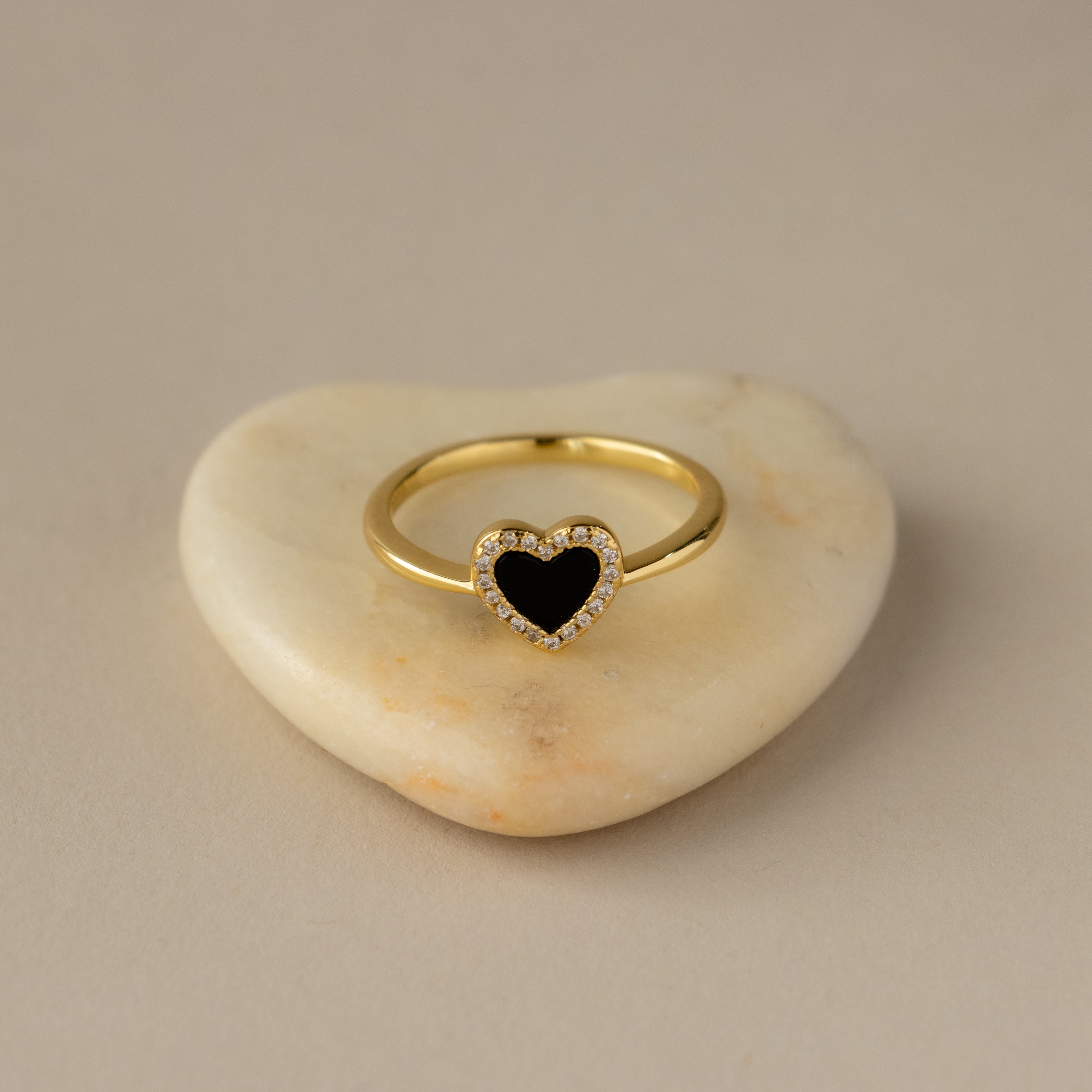 The Black Pave Heart Ring features a gold band with a black heart-shaped stone and sparkling diamonds, displayed on a white heart-shaped stone.
