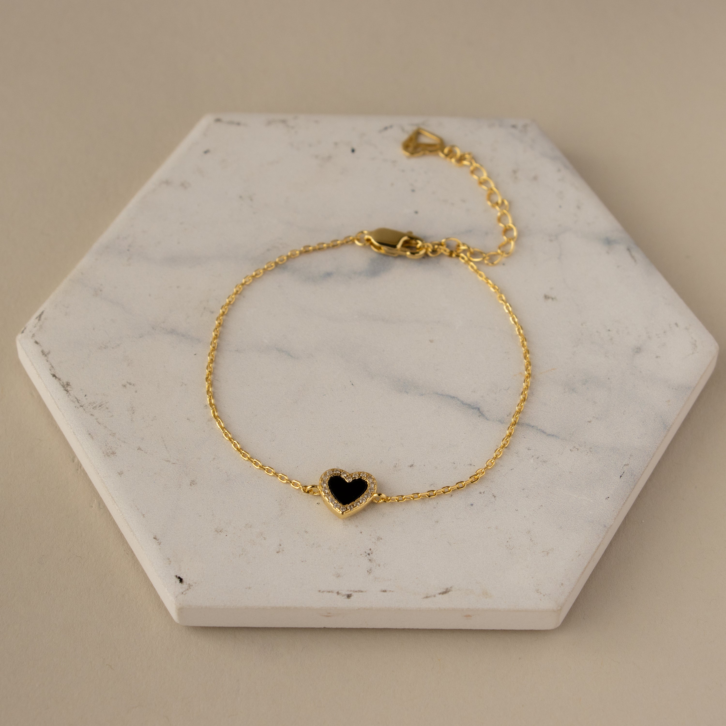 The Black Pave Heart Bracelet, featuring a gold chain and black heart charm, is displayed on a white hexagonal marble slab.