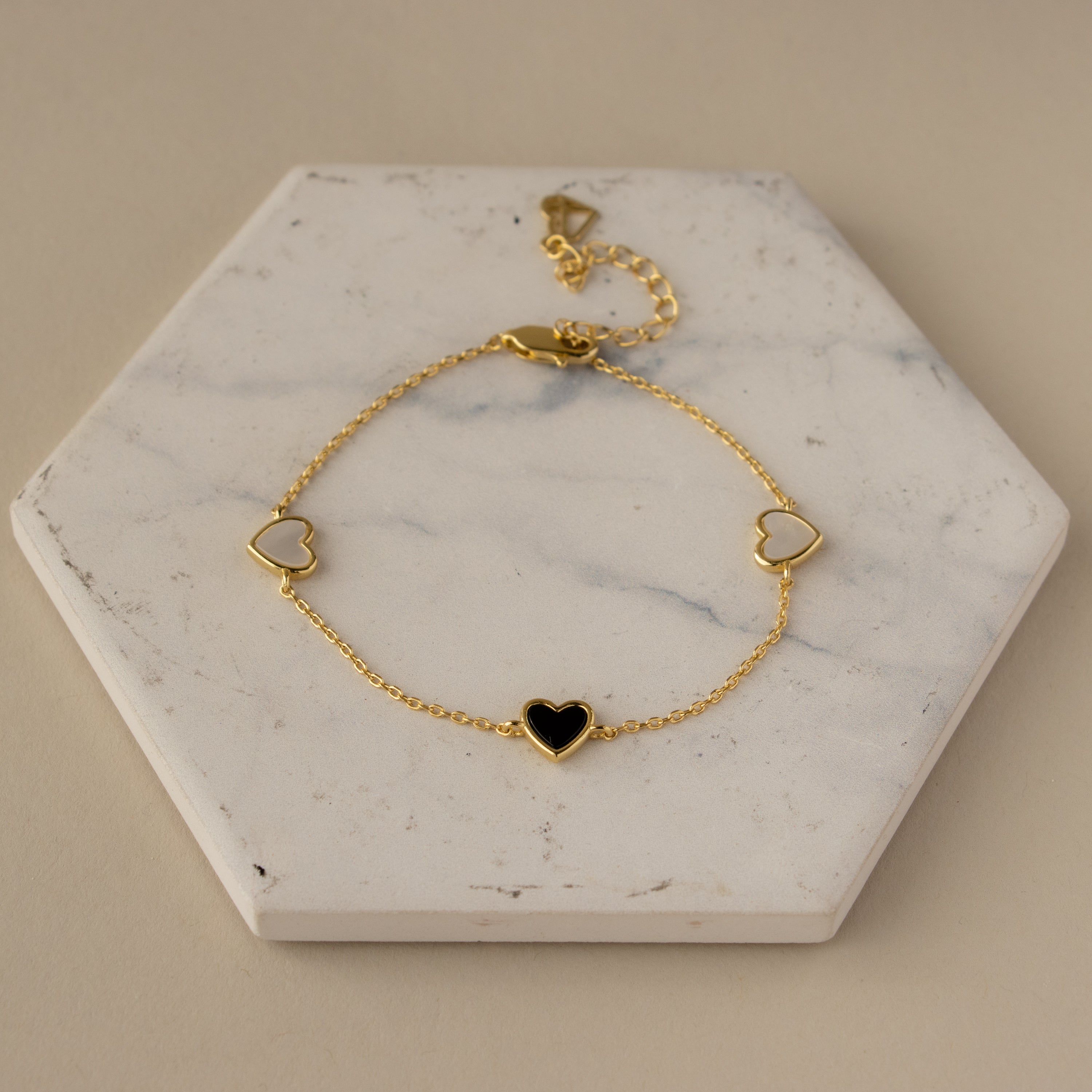 Mother of Pearl & Onyx Heart Charm Bracelet featuring three heart charms, displayed on a white hexagonal marble slab.