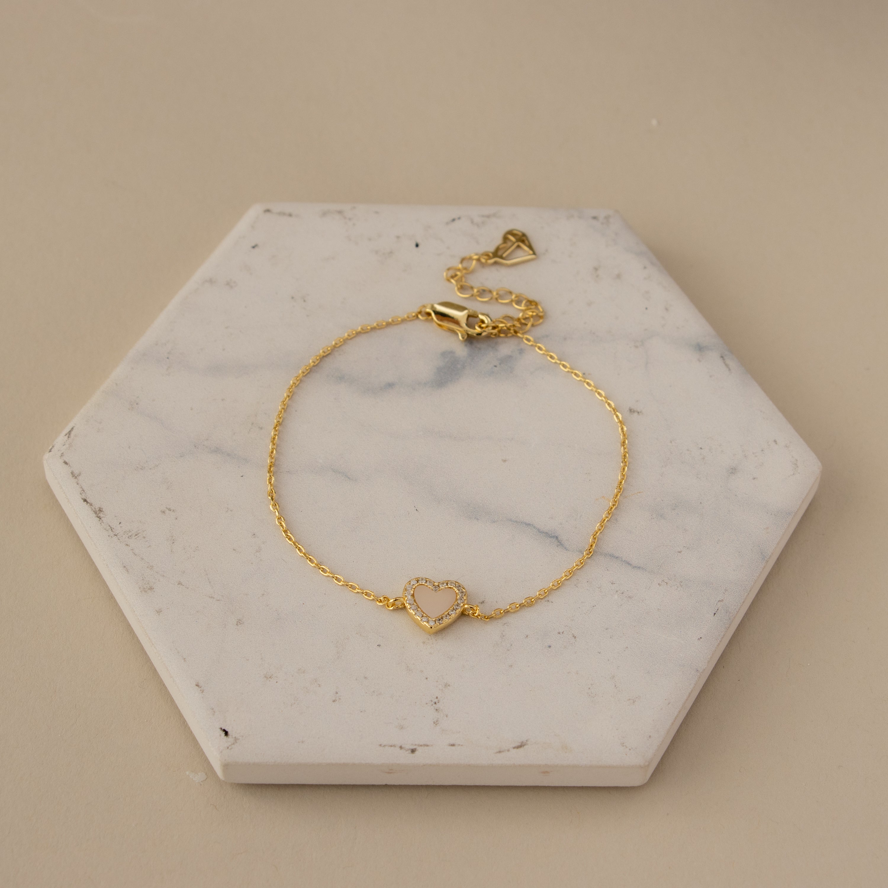 The Pink Pave Heart Bracelet, featuring a heart-shaped charm, is displayed on a white hexagonal marble slab.
