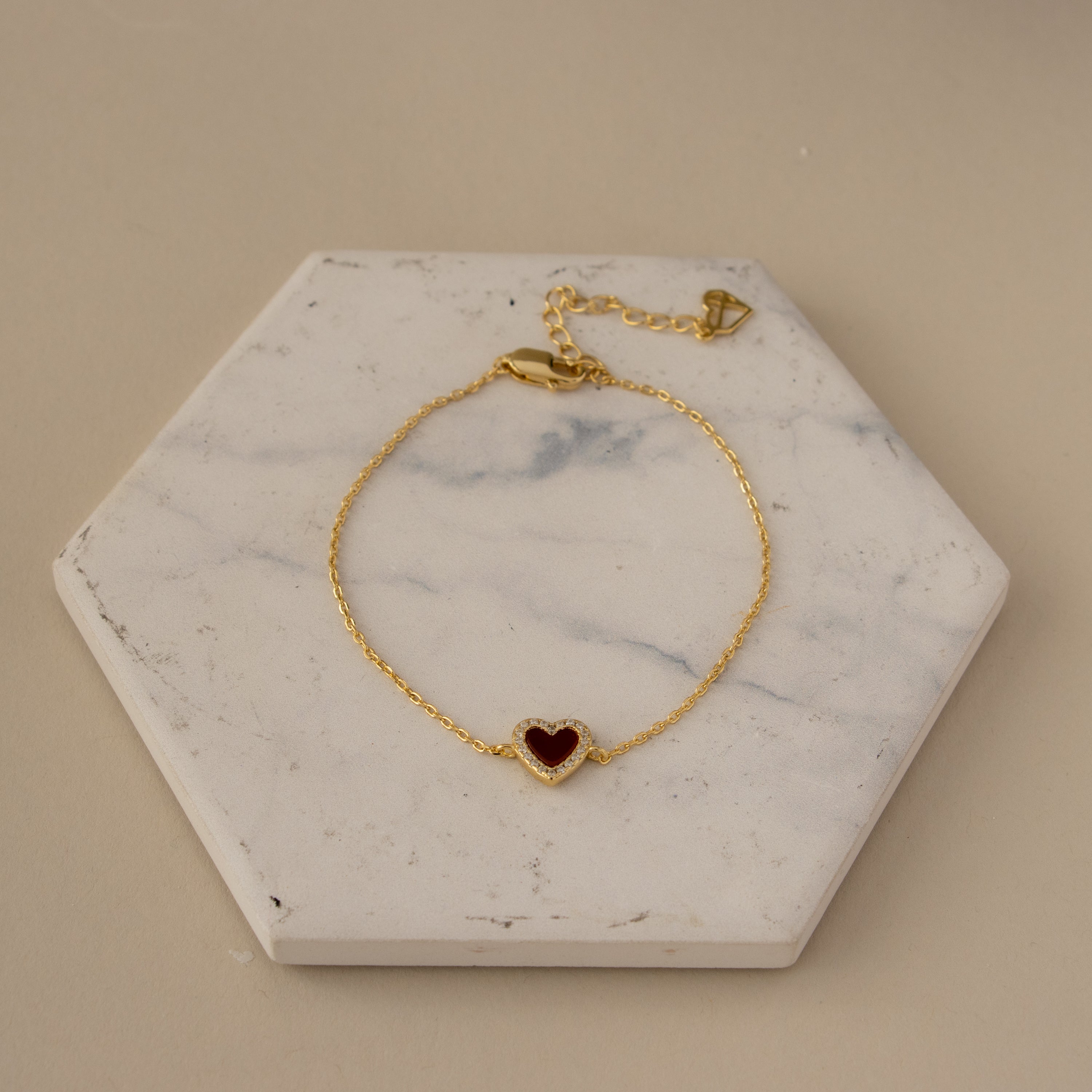 Red Agate Pave Heart Bracelet featuring a gold chain and red heart charm, showcased on a hexagonal white marble slab.
