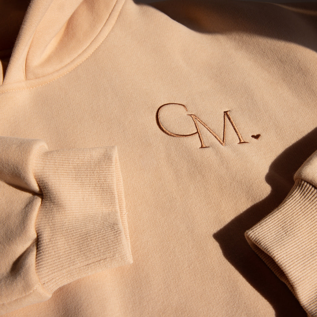 The Oversized CM Hoodie in beige features “CM.” and a small heart embroidered on the chest. Shown in natural light, this Caitlyn Minimalist plush hoodie offers effortless style and cozy comfort.