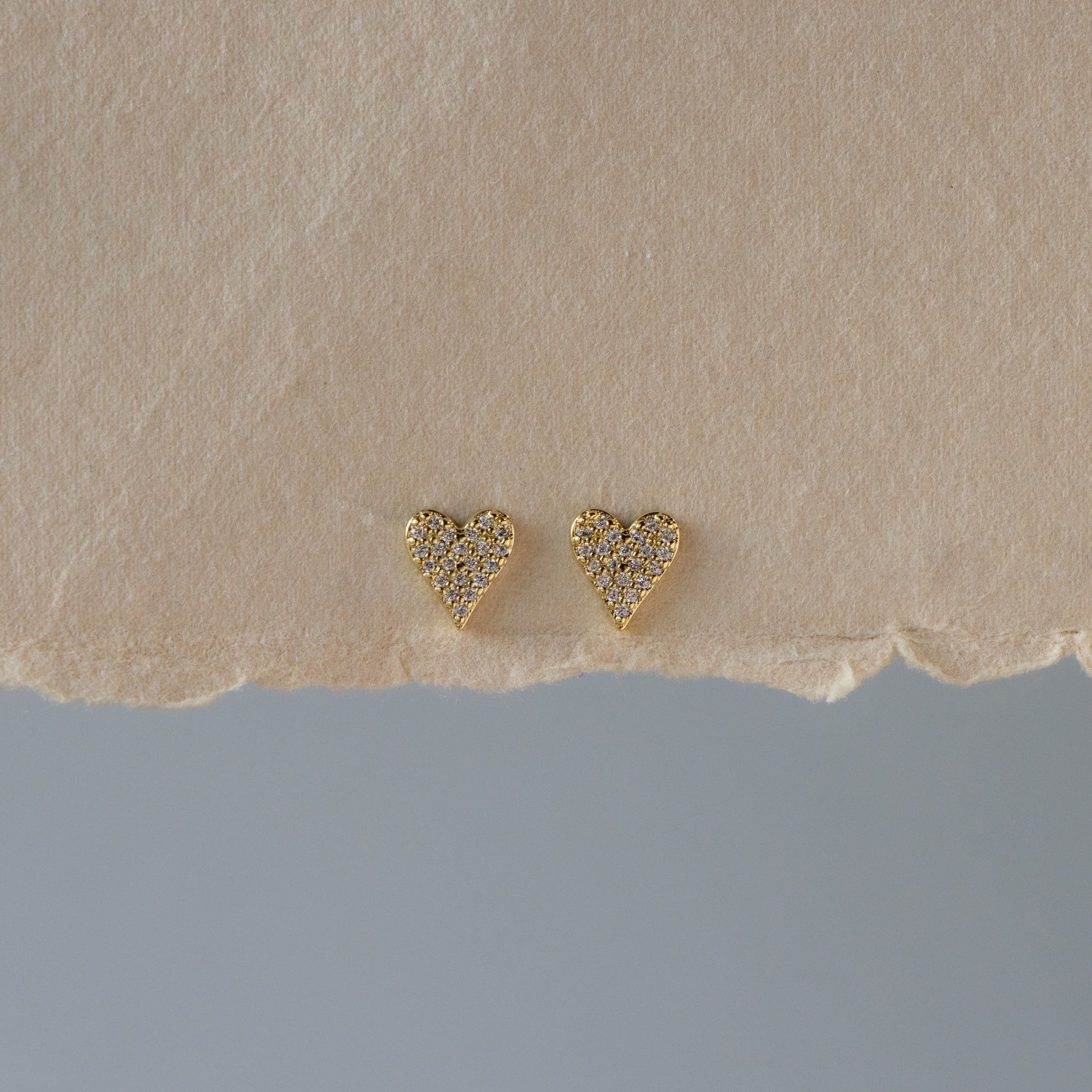 Gold Pave Heart Studs featuring small rhinestones, displayed on a textured beige surface.