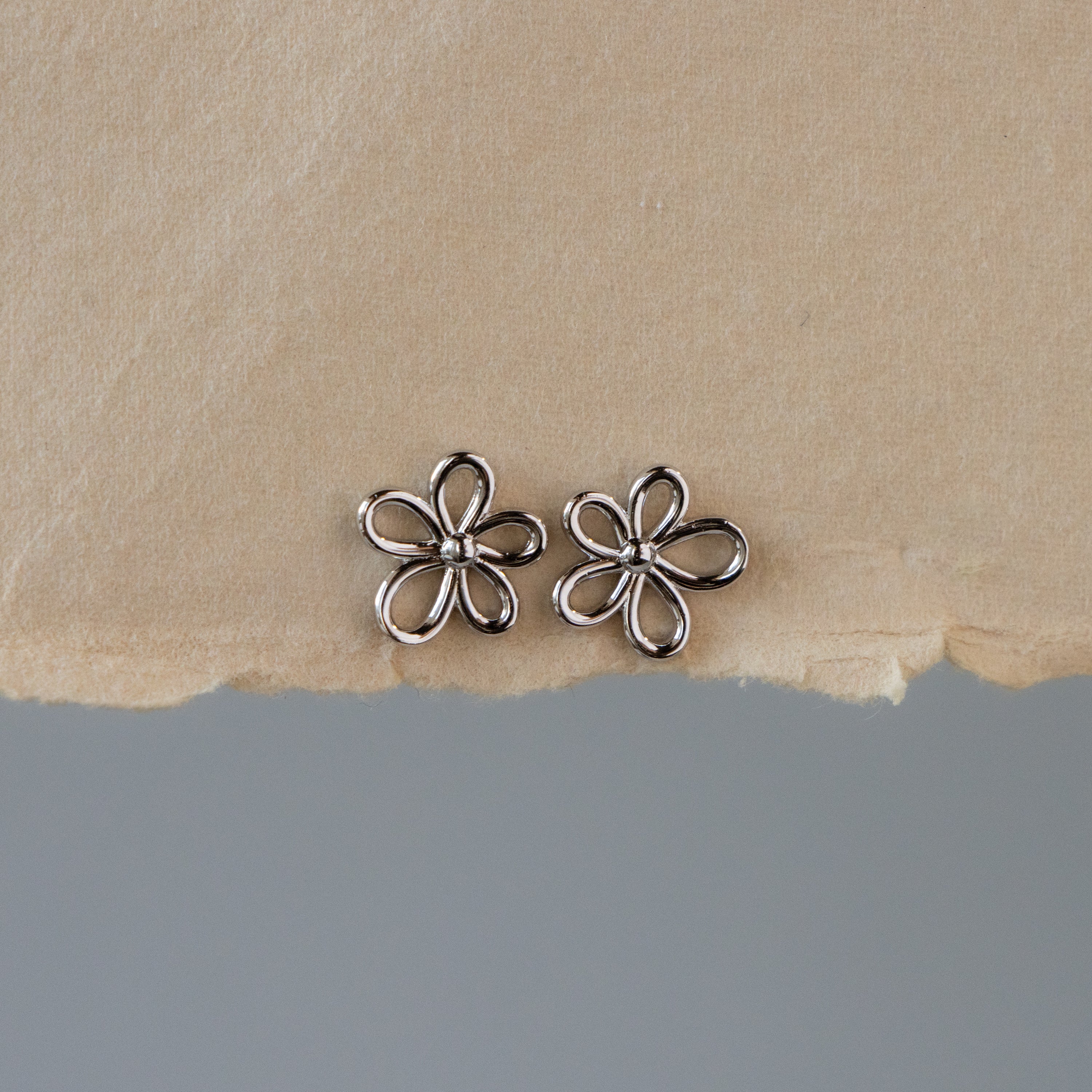 The Silver Skinny Flower Studs are displayed on a textured beige and gray background.