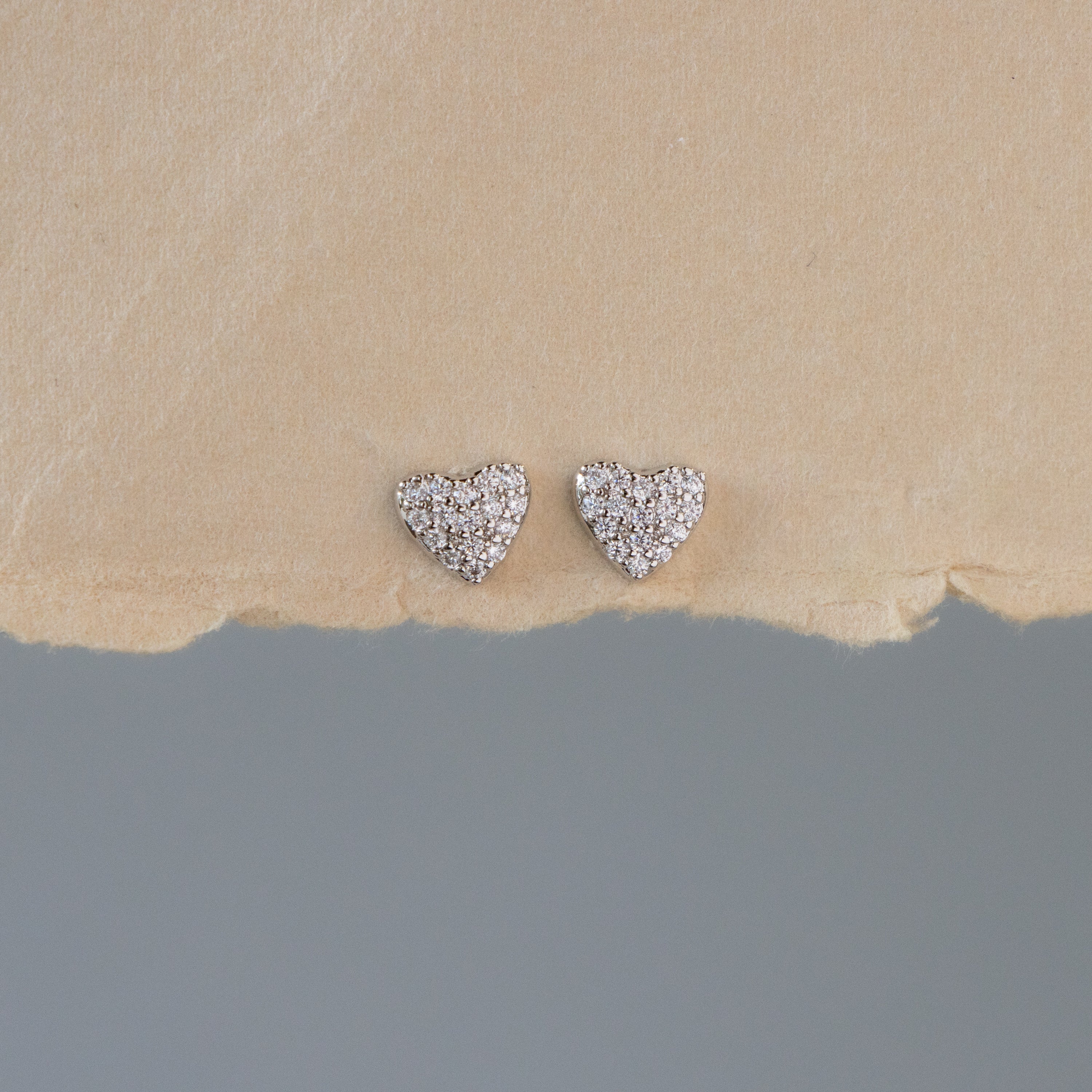 The Silver Pave Heart Studs are small, heart-shaped silver earrings adorned with sparkling stones, shown on a beige textured surface.