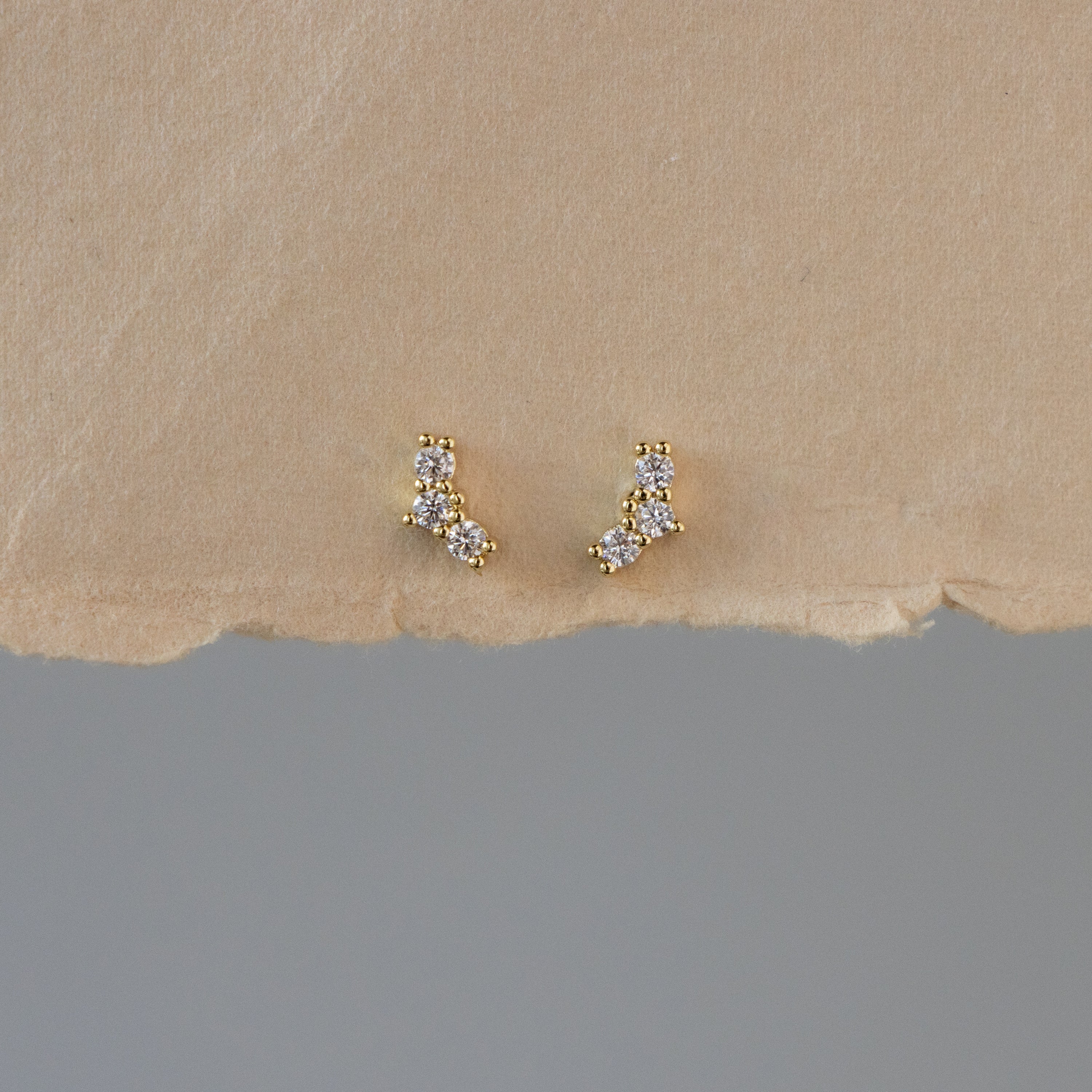 Tiny Diamond Trio Studs—gold earrings featuring small, clustered white stones—displayed on a textured neutral background.