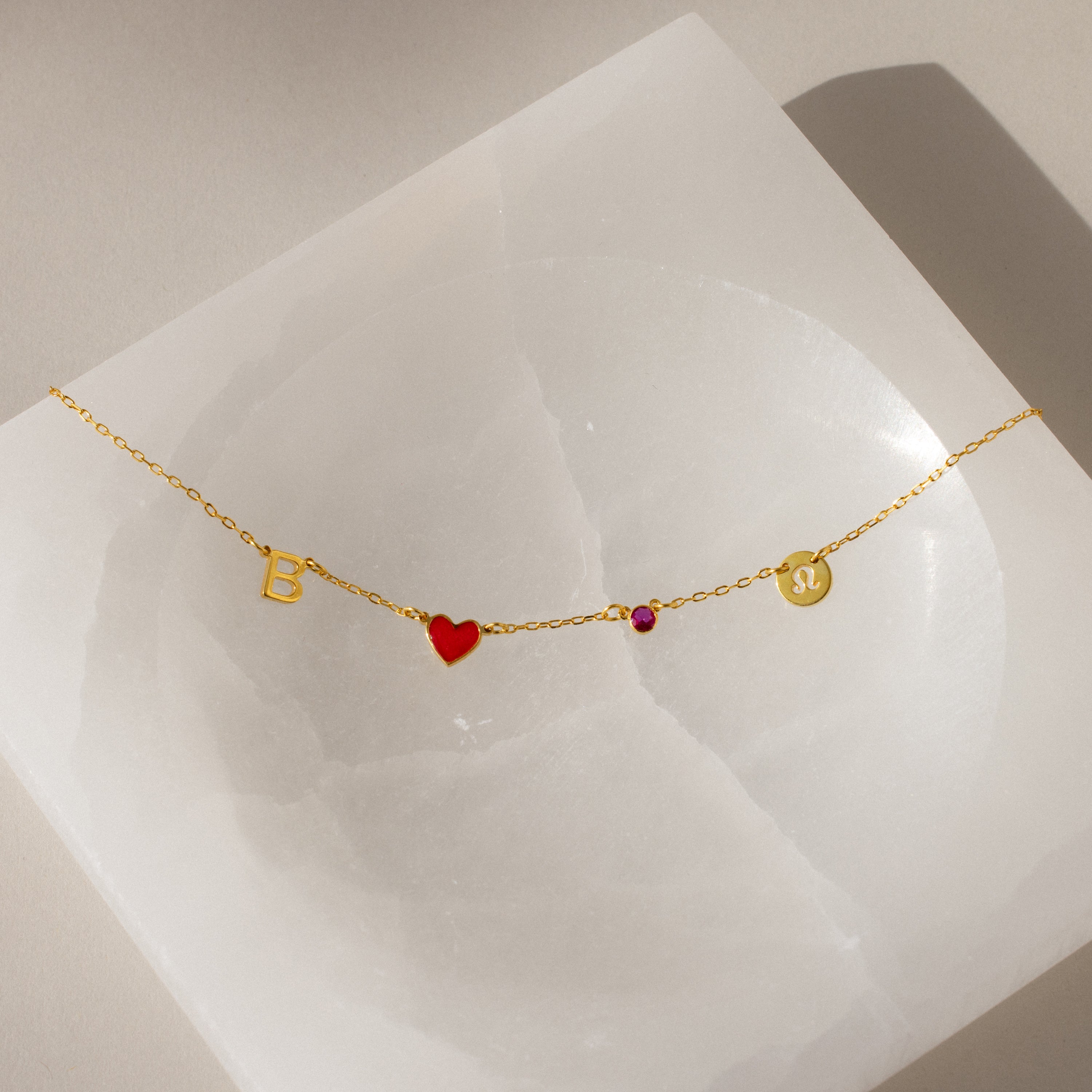 Gold bracelet with letter B, red heart, pink gem, and gold disc on a white square—perfect for pairing with the Dainty Space Charm Necklace or other customizable charm necklace styles.