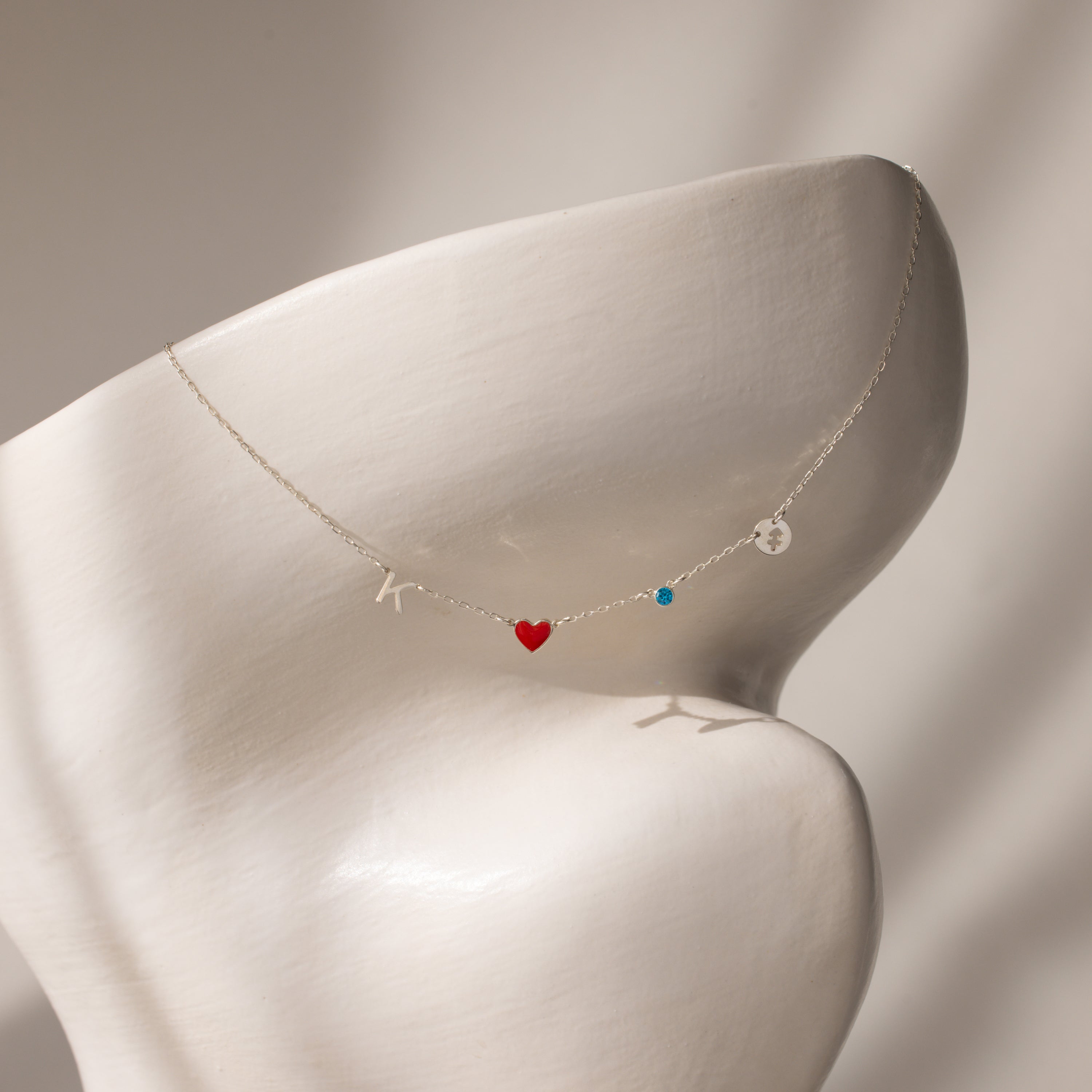 The Dainty Space Charm Necklace in Sterling Silver features a customizable K, red heart, blue bead, and round charm—ideal for anyone seeking personalized jewelry with a delicate touch.