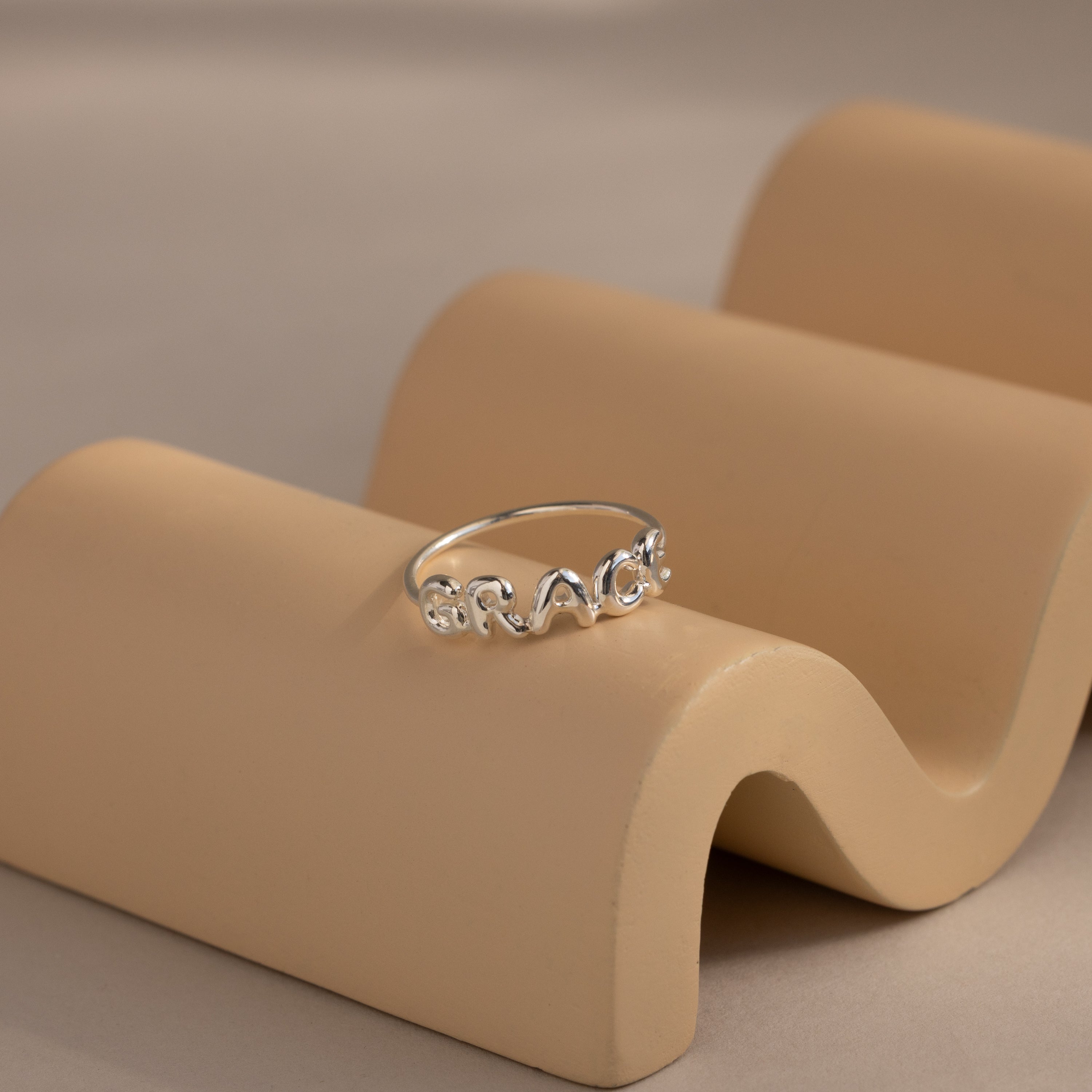 Silver bubble letter ring with the name "GRACE" resting on a neutral-tone wavy ceramic dish.