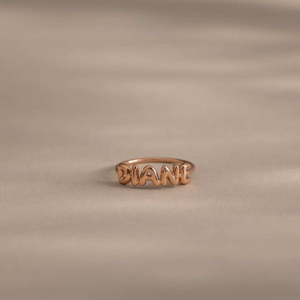 Up close of a dainty rose gold bubble letter ring with the name "DIANE" displayed on a neutral surface.