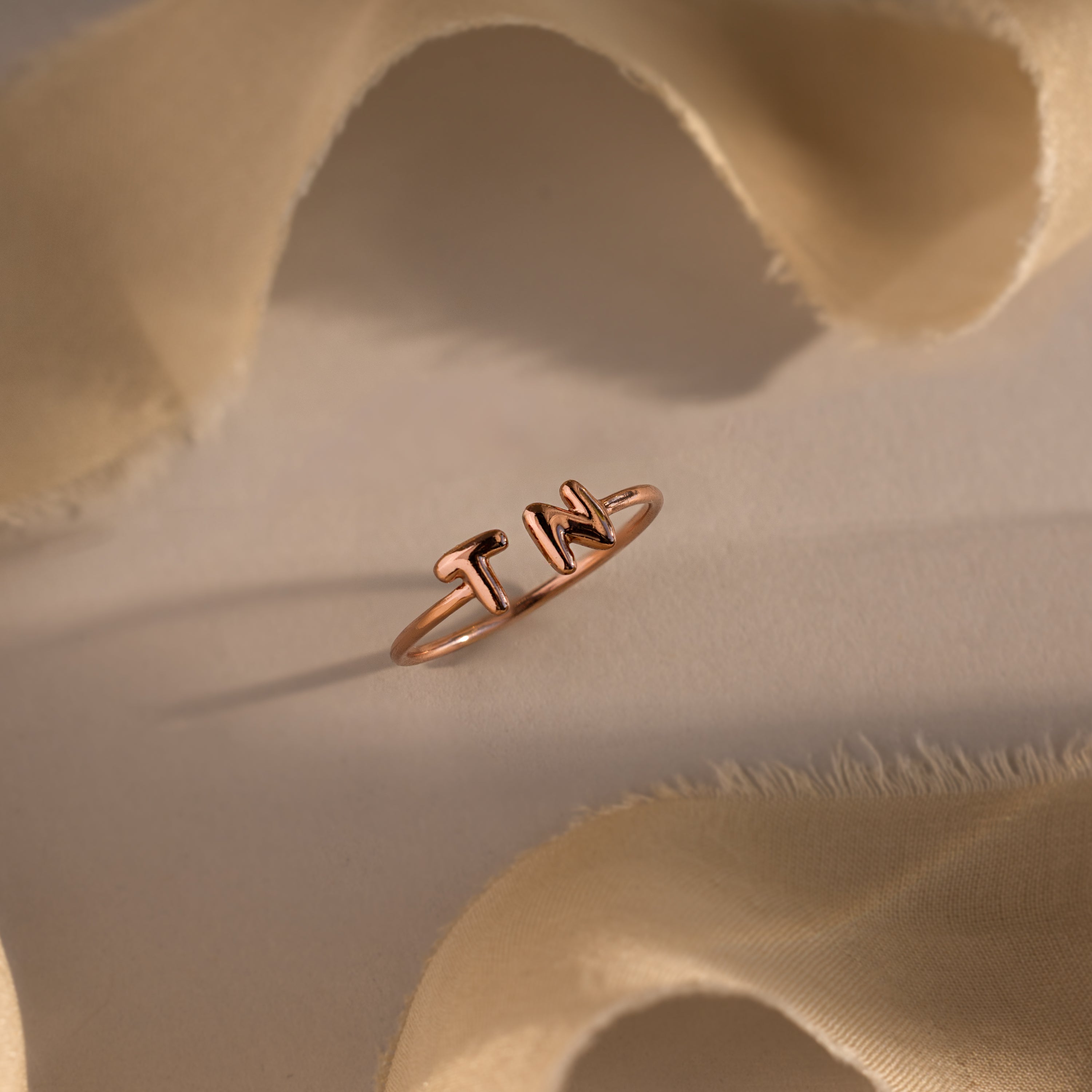 Close-up of a rose gold custom initial ring with bubble letters T and N, styled on a soft beige background with flowing fabric.