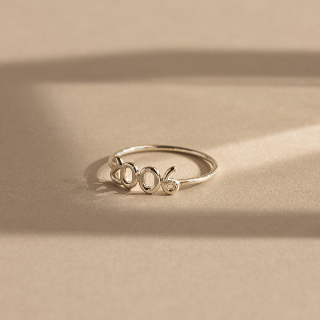 A silver “2006” Bubble Numbers Ring on a beige background. The ring has rounded bubble numbers with a smooth and shiny finish.