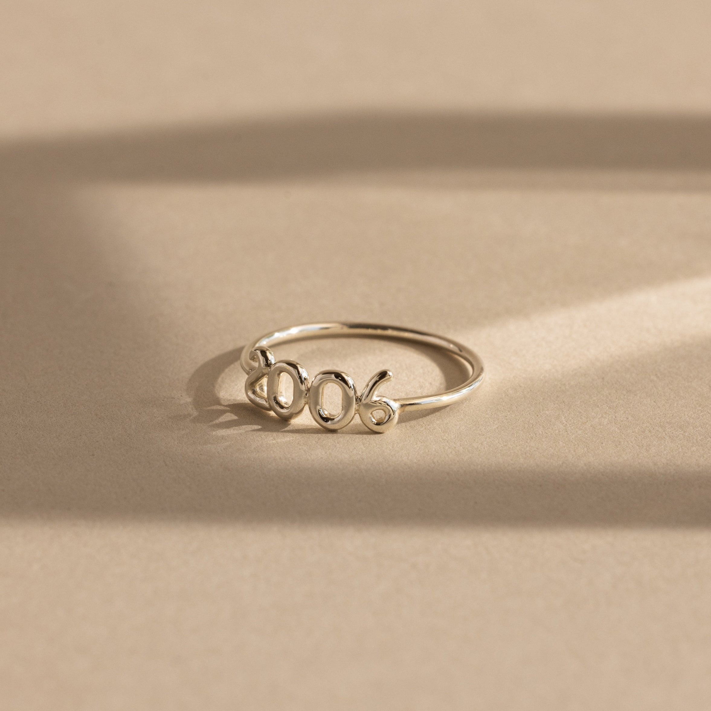 A silver “2006” Bubble Numbers Ring on a beige background. The ring has rounded bubble numbers with a smooth and shiny finish.