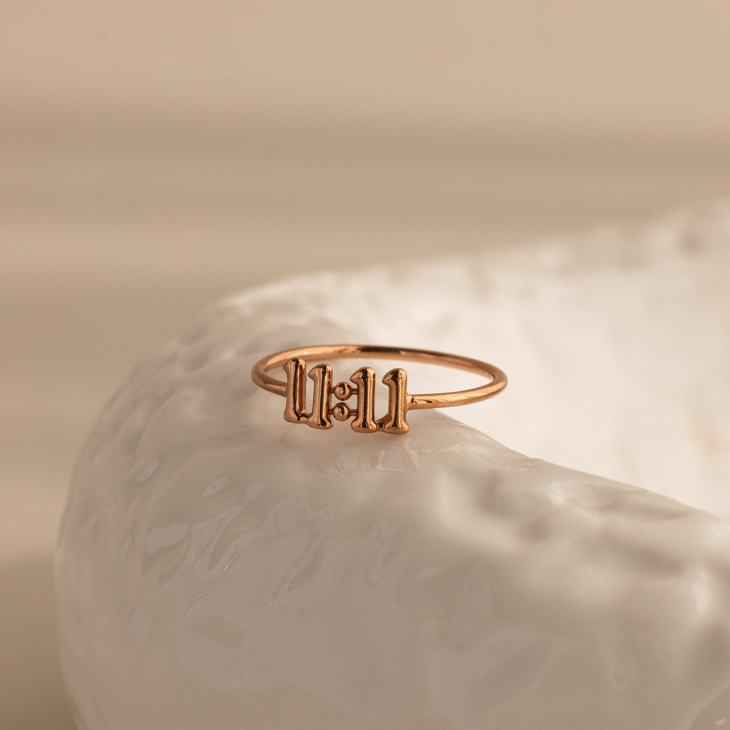 A close-up of a rose gold Bubble Numbers Ring featuring the number “11 11” in bold, rounded numerals, styled on the edge of a glossy, textured white ceramic surface with soft beige lighting.
