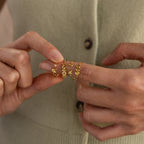 A close-up of hands displaying a stack of gold rings with bold bubble-style letters, including initials "A", "K", "J", numbers "444", and names like "MOMMY", worn and held on a single finger against a soft sage knit top, with one hand gently lifting a ring featuring a single round gemstone.