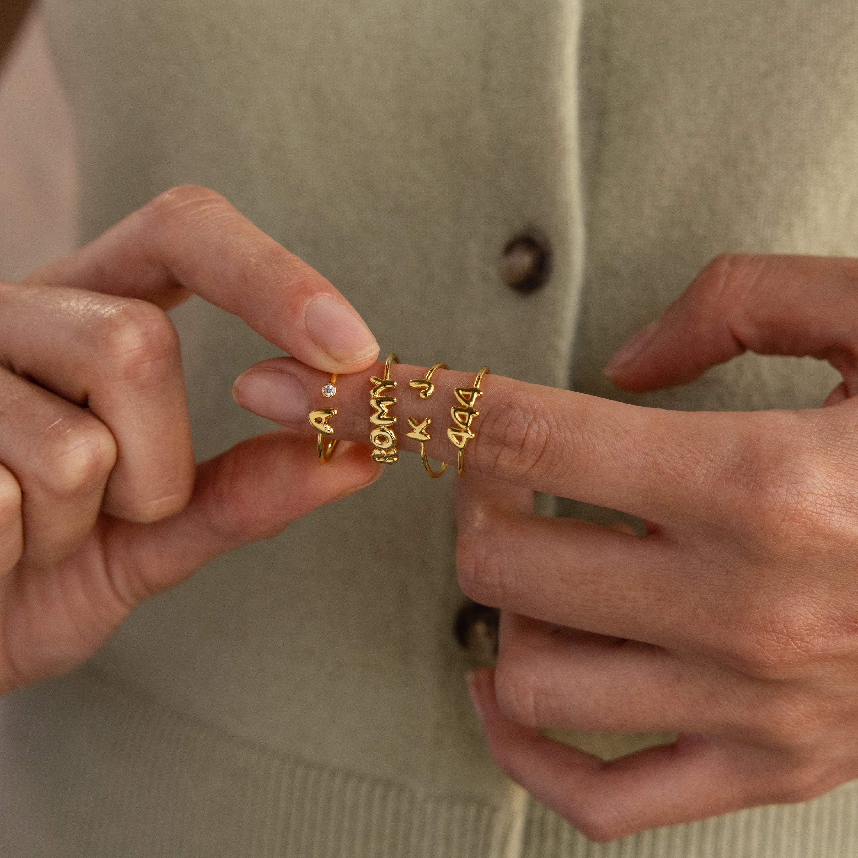 A close-up of hands displaying a stack of gold rings with bold bubble-style letters, including initials "A", "K", "J", numbers "444", and names like "MOMMY", worn and held on a single finger against a soft sage knit top, with one hand gently lifting a ring featuring a single round gemstone.