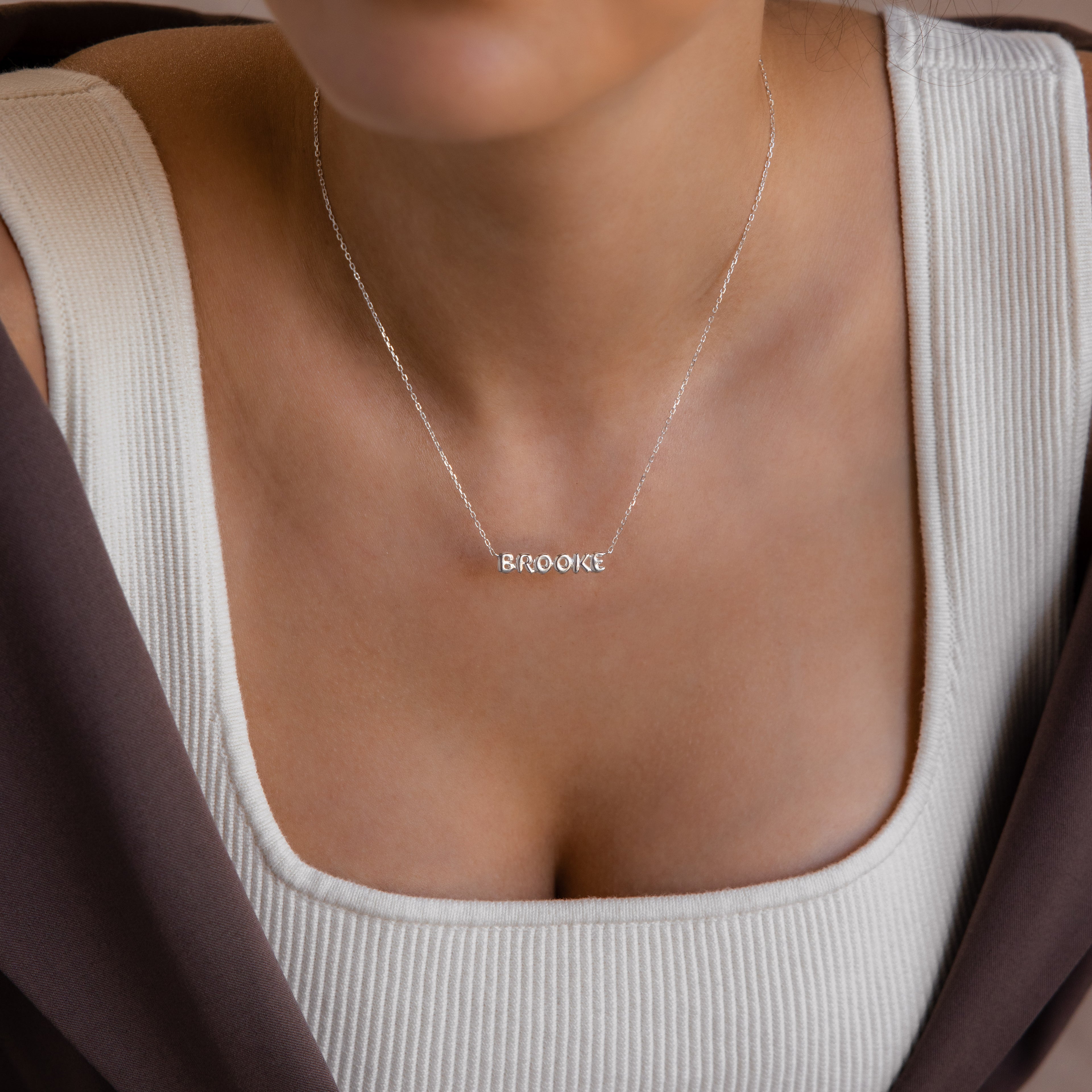 A woman wearing a Tiny Bubble Name Necklace personalized with BROOKE, dressed in a white top and brown jacket.