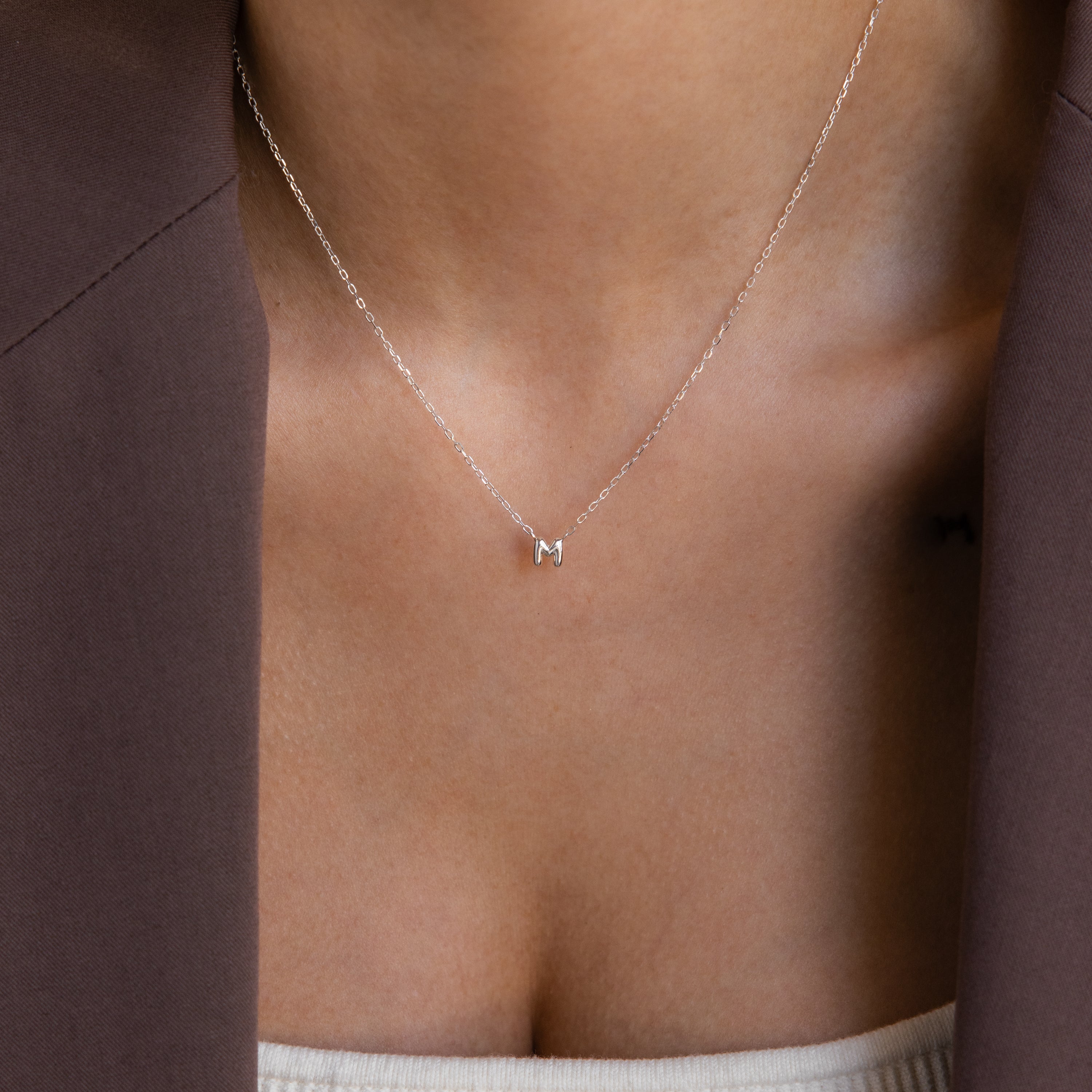 Model wears a dainty silver bubble letter M charm necklace.