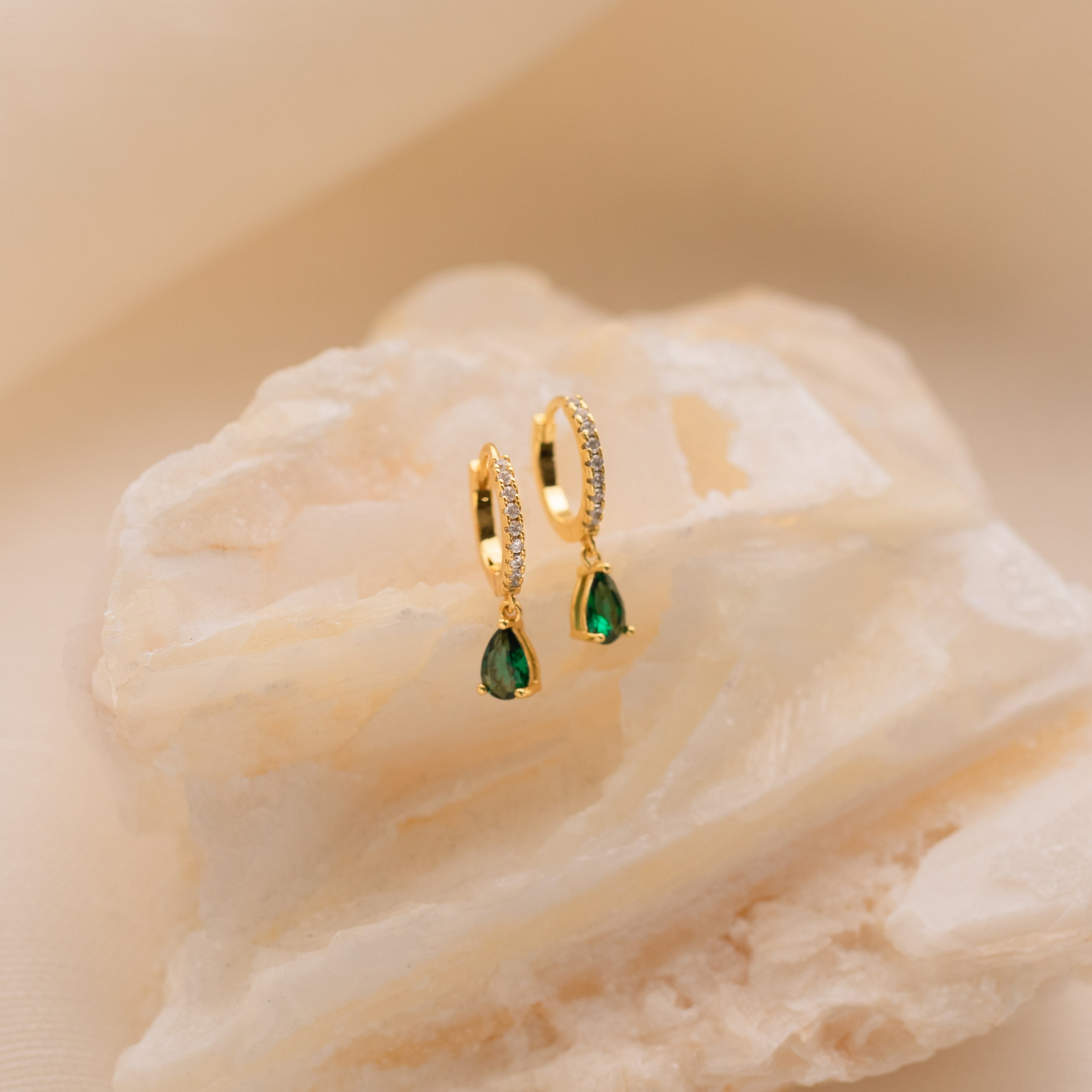 Gold hoop earrings with green birthstone teardrops, shown on a textured white stone background, highlight the elegance of Pave Birthstone Drop Huggies.