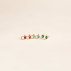 A gold ring with pink, blue, and green gemstones is displayed on a light background next to Raindrop Peridot Studs.