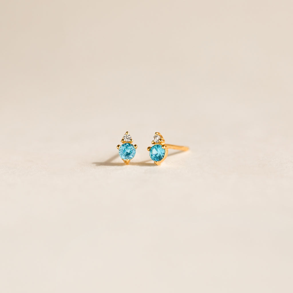 Raindrop Blue Zircon Studs, featuring gold dainty earrings with blue gemstones and small white stones, are displayed on a light, neutral background.