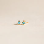 Raindrop Blue Zircon Studs, featuring gold dainty earrings with blue gemstones and small white stones, are displayed on a light, neutral background.