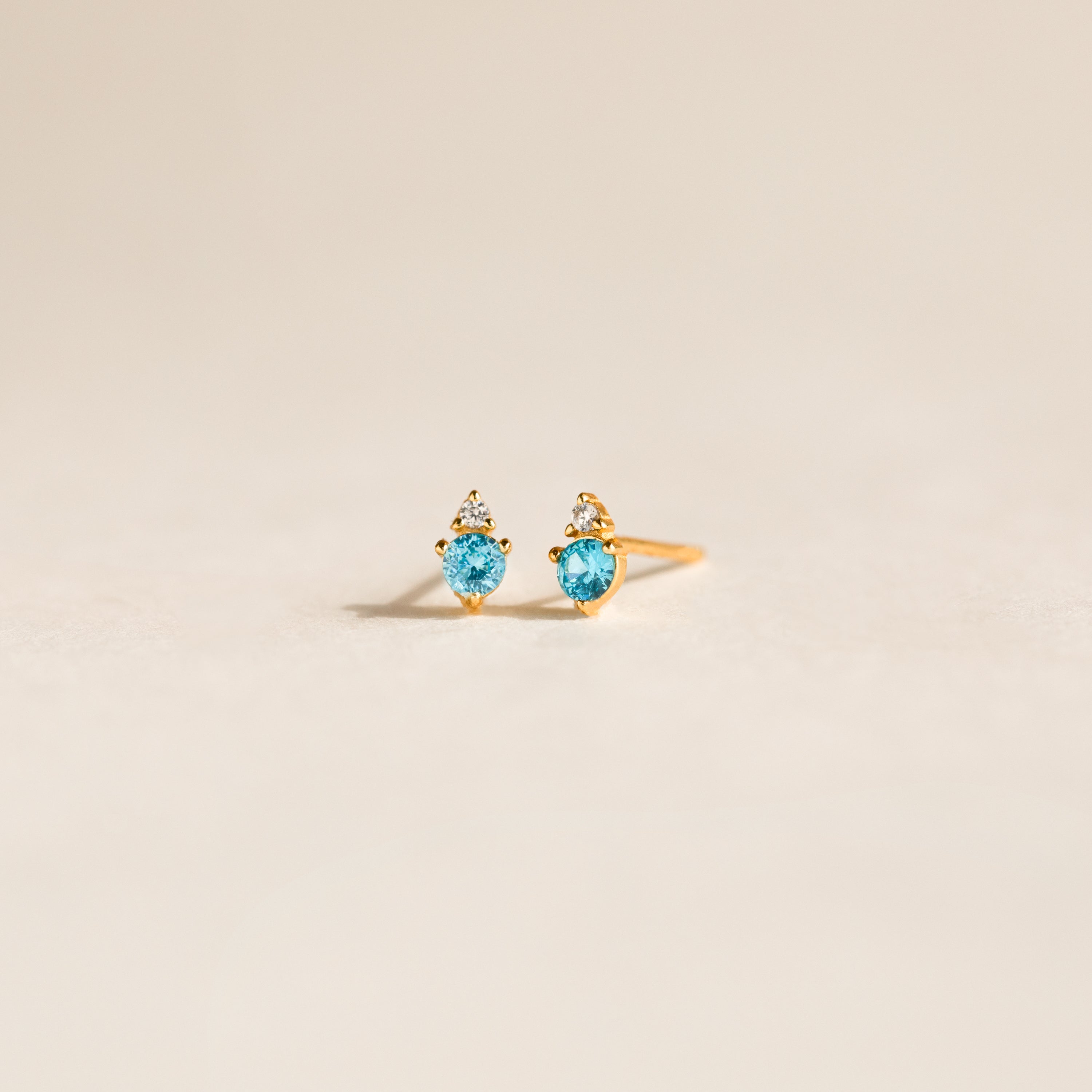 Raindrop Blue Zircon Studs, featuring gold dainty earrings with blue gemstones and small white stones, are displayed on a light, neutral background.