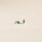 Raindrop Blue Zircon Studs feature gold settings with small blue zircons and clear accents, creating dainty, elegant birthstone earrings on a light beige background.