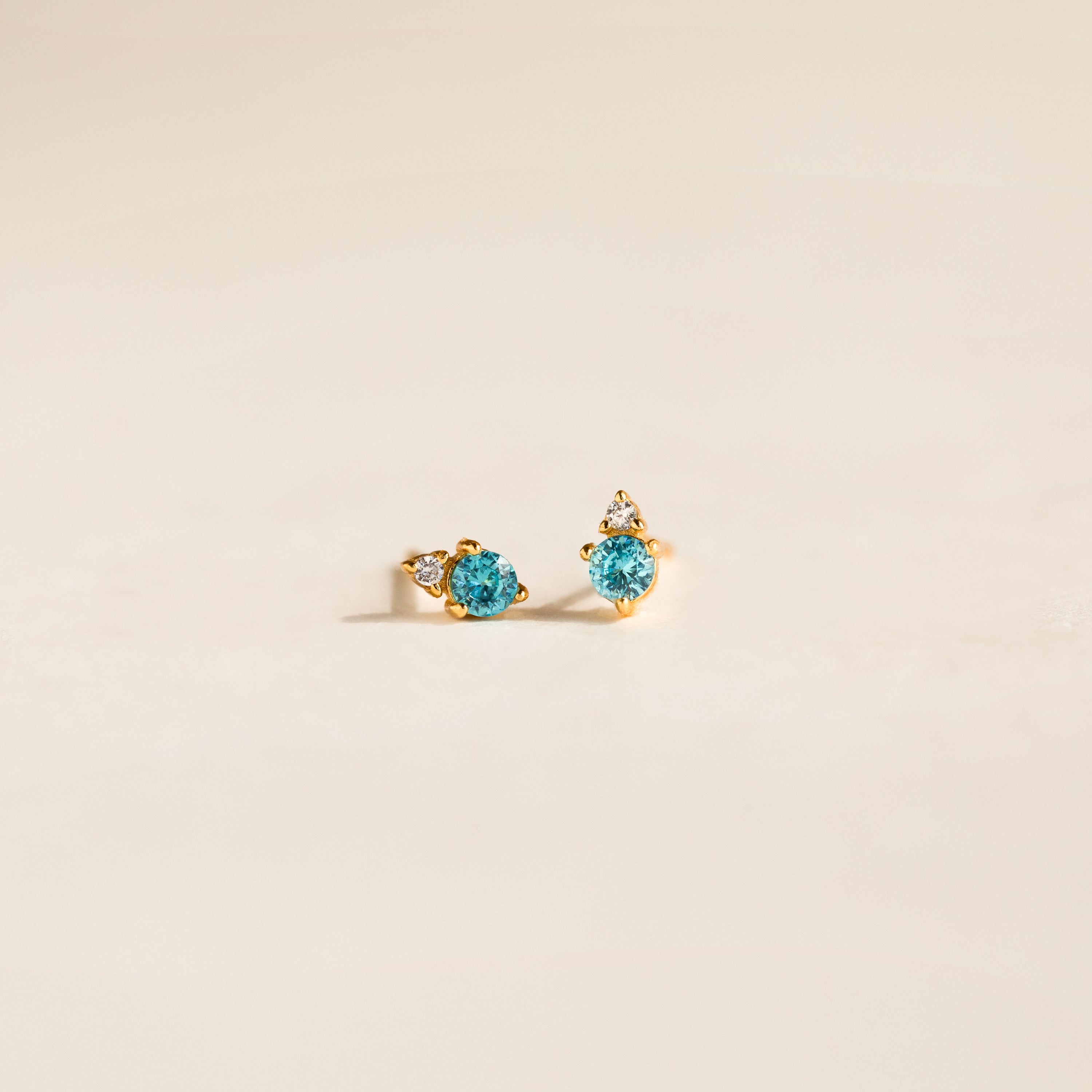 Raindrop Blue Zircon Studs feature gold settings with small blue zircons and clear accents, creating dainty, elegant birthstone earrings on a light beige background.