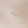 The Bella Blue Zircon Studs feature three sparkling blue gemstones on each gold earring, displayed on soft beige fabric—an ideal gift for mothers and daughters.
