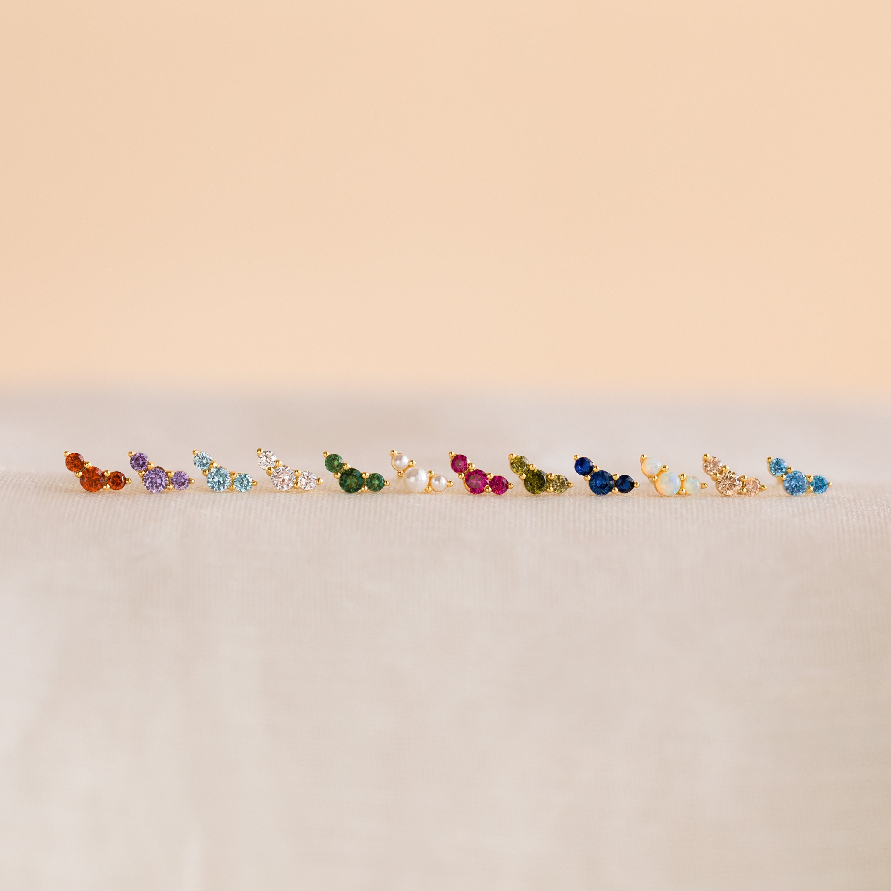 Bella Birthstone Studs—vivid, petite earrings arranged on a soft, neutral surface with a beige backdrop—make perfect personalized jewelry and a thoughtful gift for mothers and daughters.