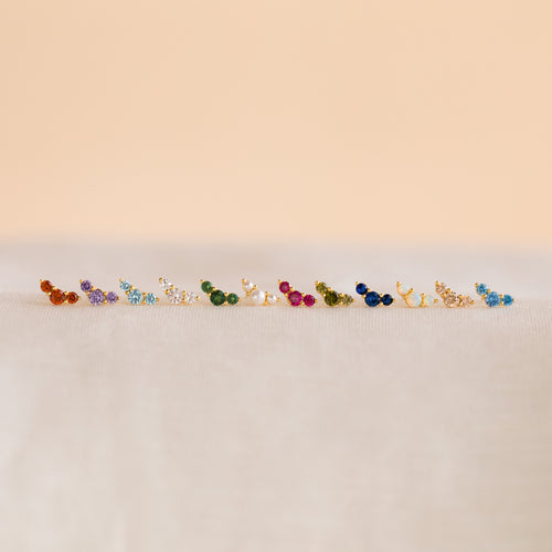 Bella Birthstone Studs—vivid, petite earrings arranged on a soft, neutral surface with a beige backdrop—make perfect personalized jewelry and a thoughtful gift for mothers and daughters.