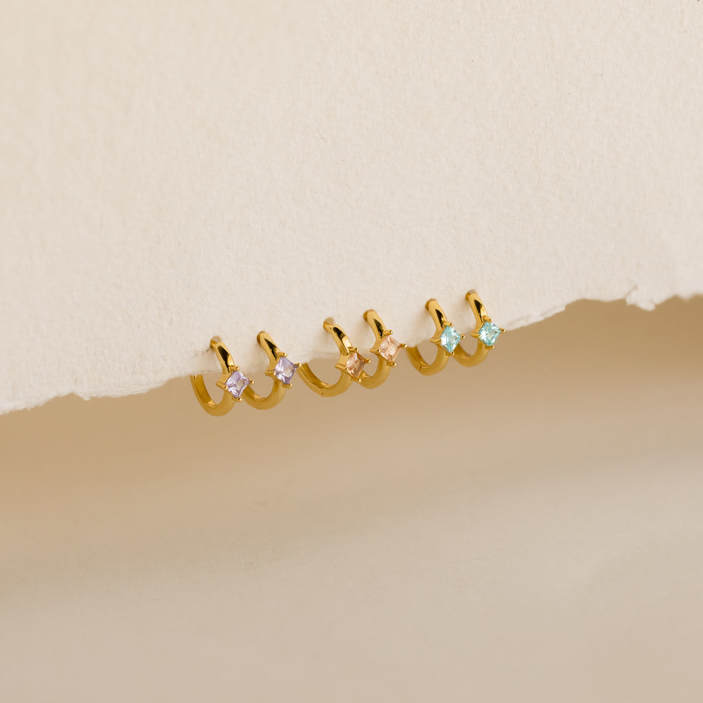 A row of pairs of small gold hoop earrings, each adorned with a square-cut gemstone in 3 colors: light purple, blush pink, and aqua blue, displayed against a textured off-white background.