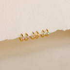 A row of pairs of small gold hoop earrings, each adorned with a square-cut gemstone in 3 colors: light purple, blush pink, and aqua blue, displayed against a textured off-white background.