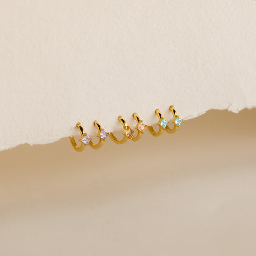 A row of pairs of small gold hoop earrings, each adorned with a square-cut gemstone in 3 colors: light purple, blush pink, and aqua blue, displayed against a textured off-white background.