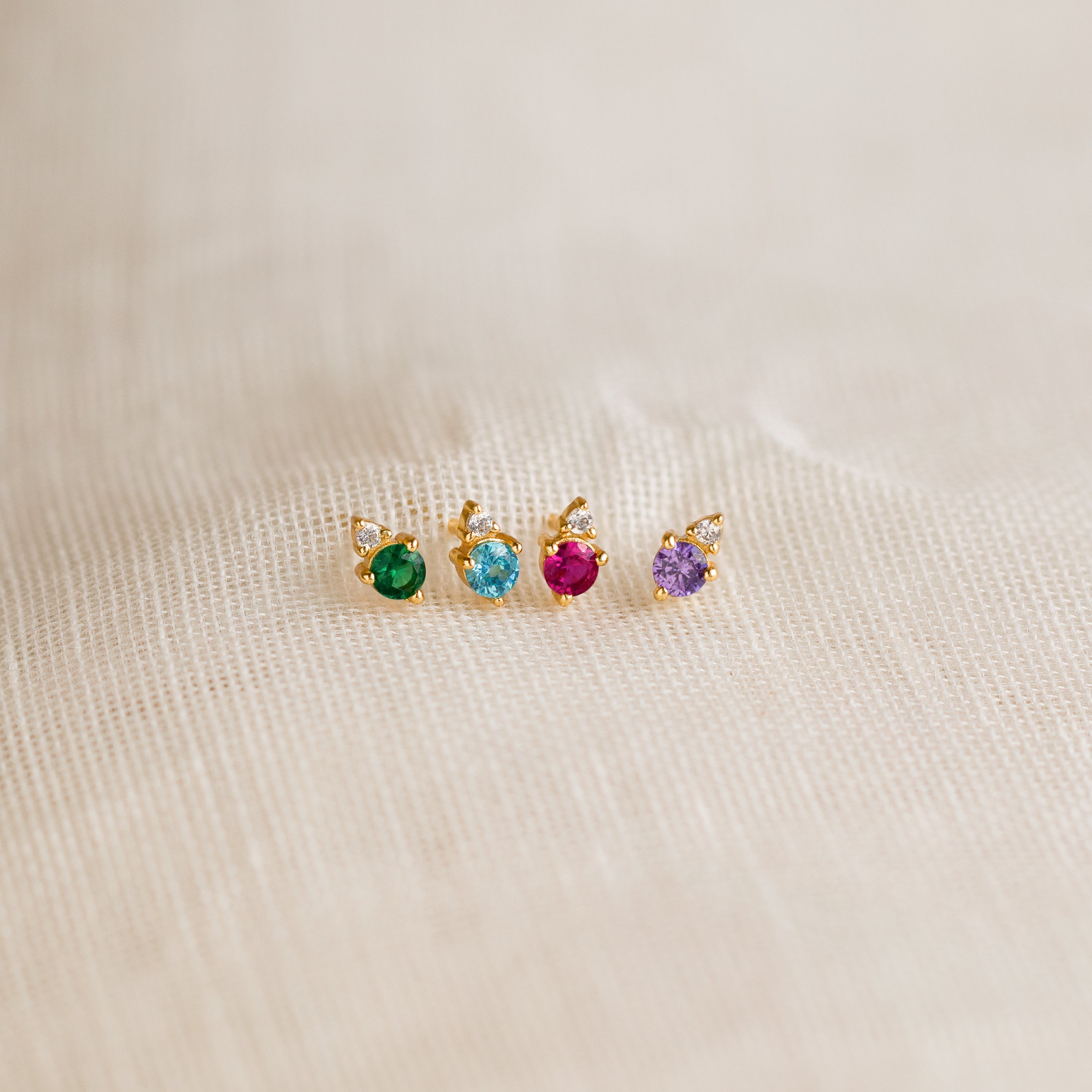 The Raindrop Emerald Studs—dainty gold earrings with green gemstones—are shown on beige textured fabric, perfect for wearing as elegant birthstone studs.