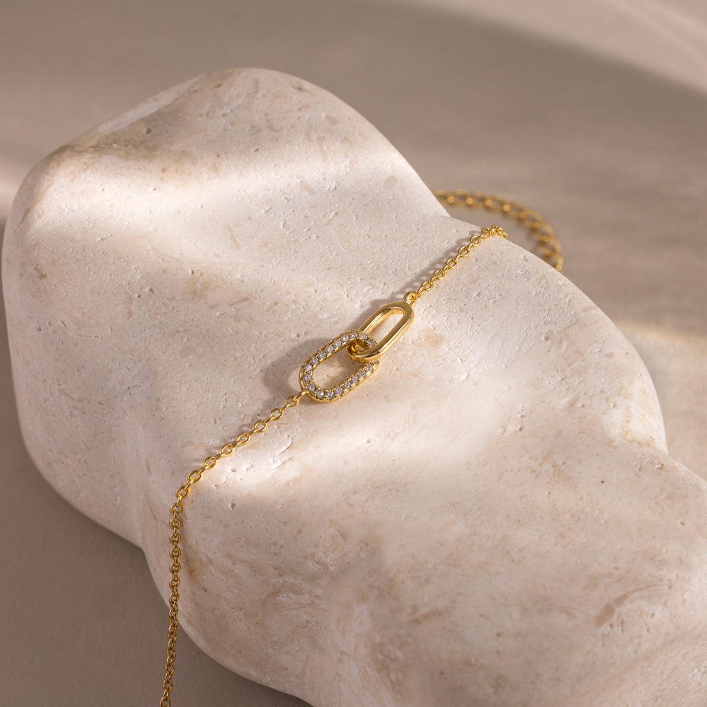 Delicate gold chain bracelet displayed on a textured stone surface, featuring an interlocking oval link design with one link encrusted in small clear stones.
